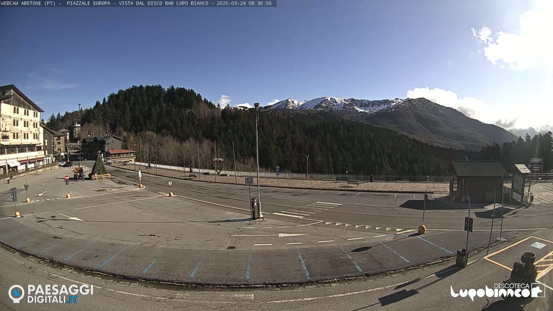 An empty parking lot and roadway sit before a snow-capped mountain range under a mostly sunny sky.
