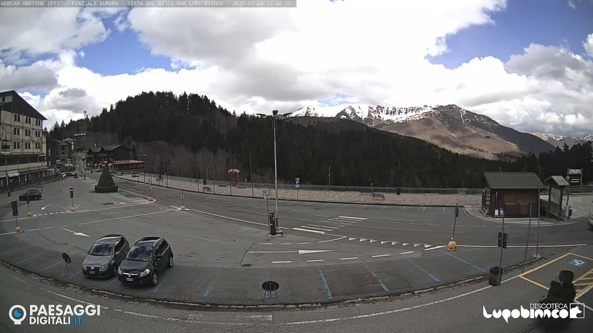 A mostly empty parking lot sits before a building and a backdrop of snow-capped mountains under a partly cloudy sky.