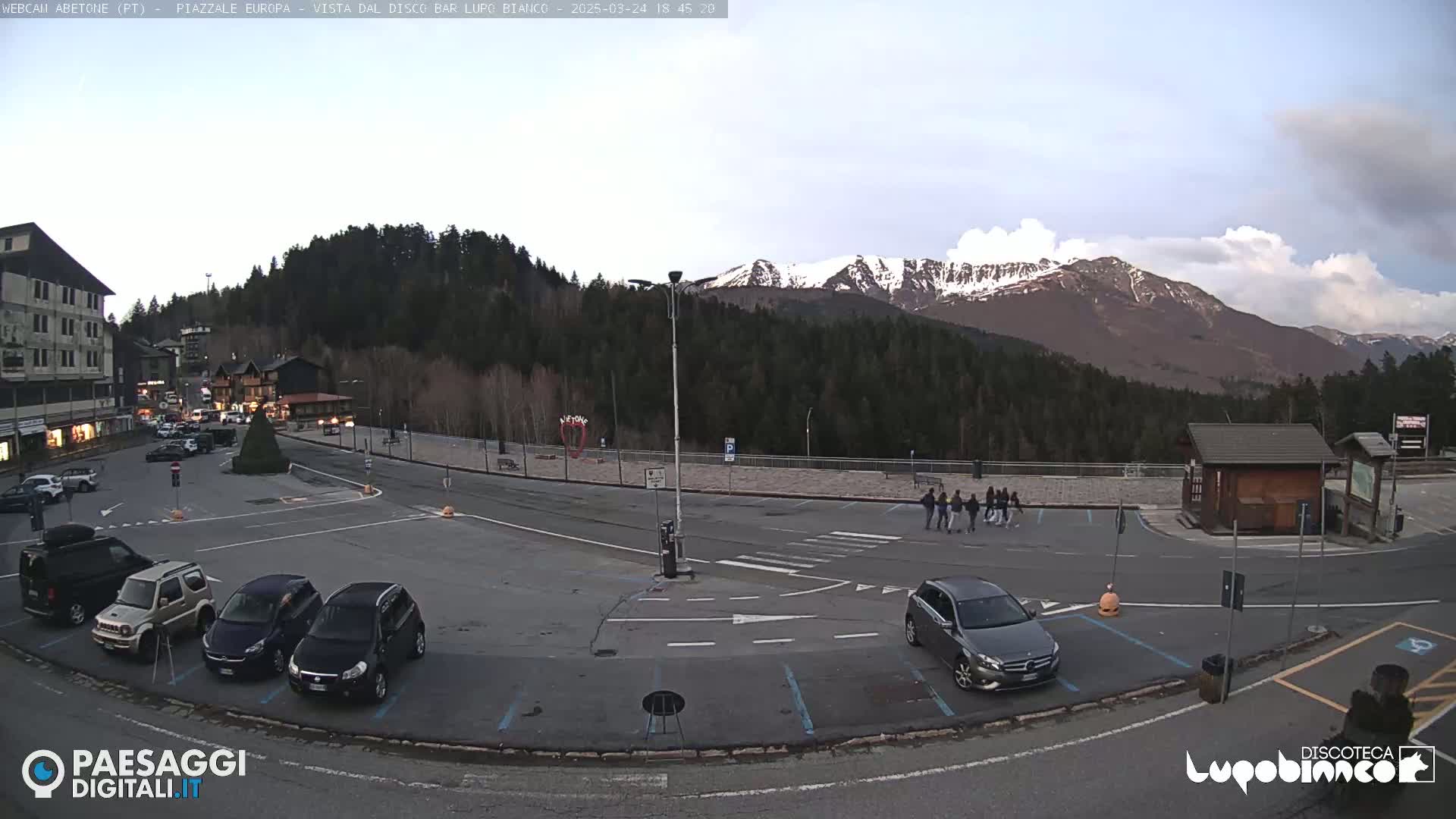 A mostly empty parking lot with several cars, a few pedestrians, and buildings in the background, overlooked by snow-capped mountains under a partly cloudy sky.