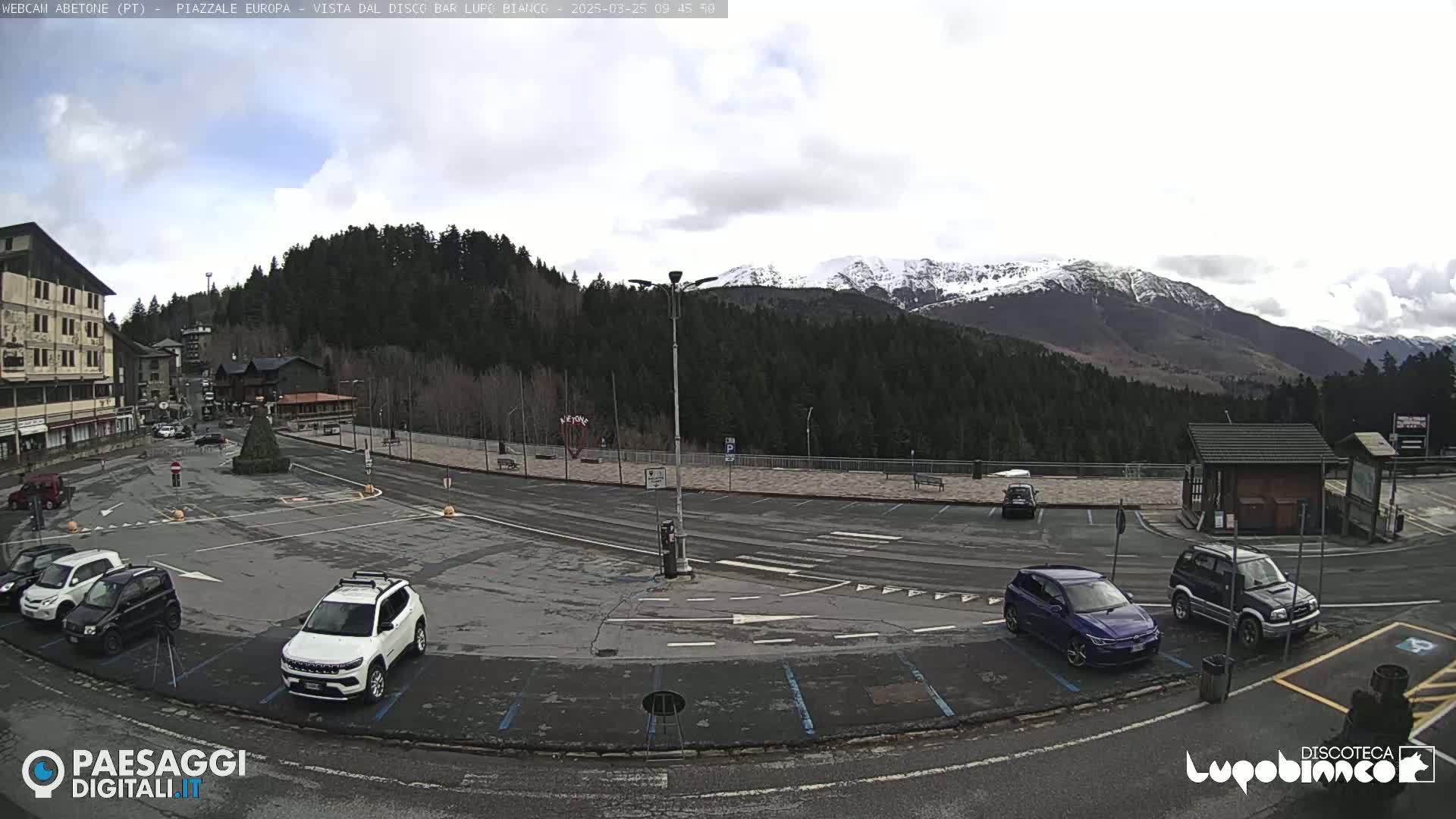 A mostly empty parking lot with several cars is situated in front of a building and a forested mountain range with snow-capped peaks under a cloudy sky.