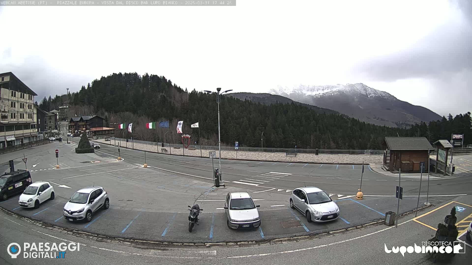 A mostly empty parking lot is situated in front of a building and overlooks a snow-capped mountain range under an overcast sky.