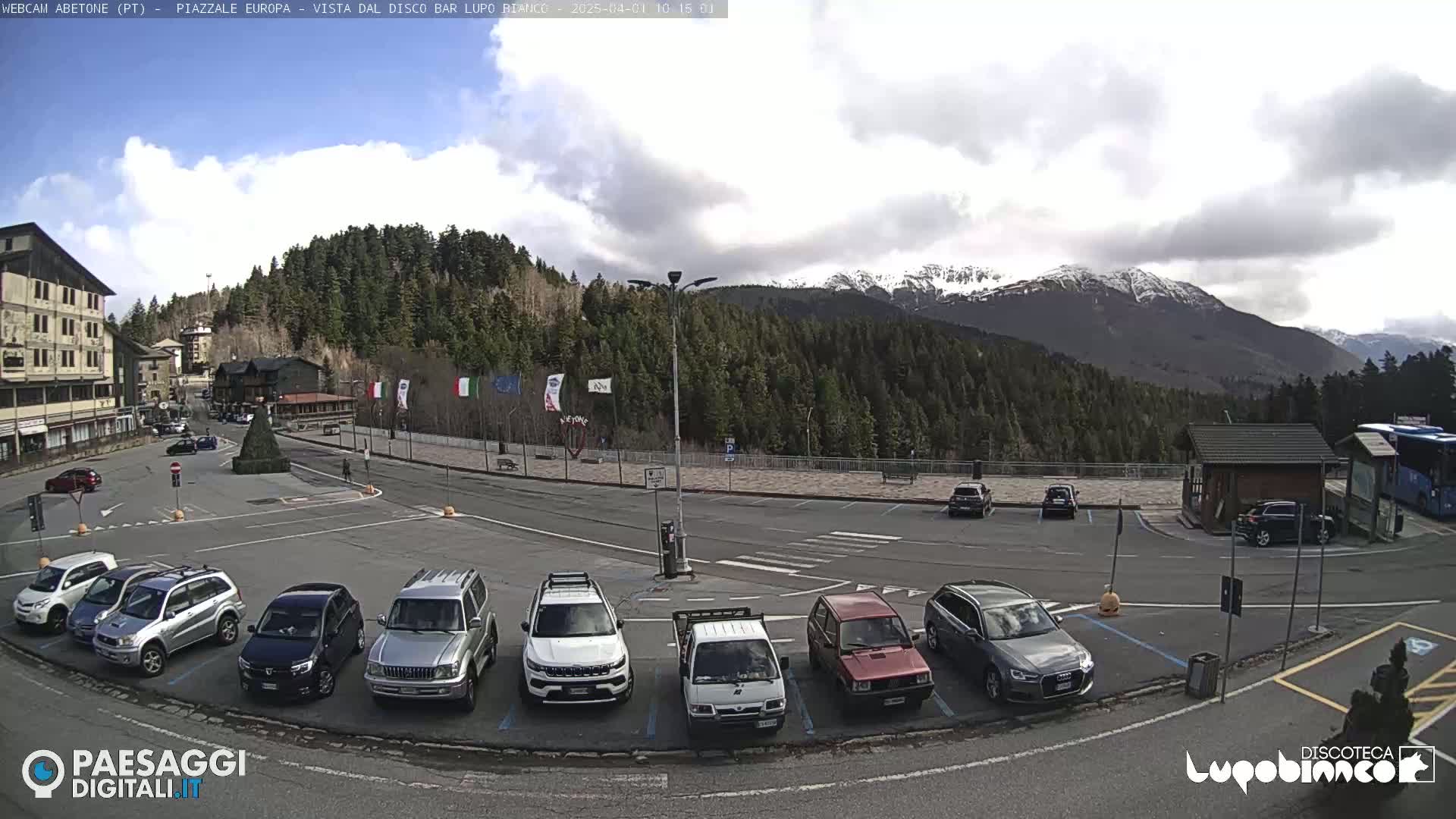 A parking lot with several cars is situated in front of a building and a wooded mountain range with snow-capped peaks under a partly cloudy sky.