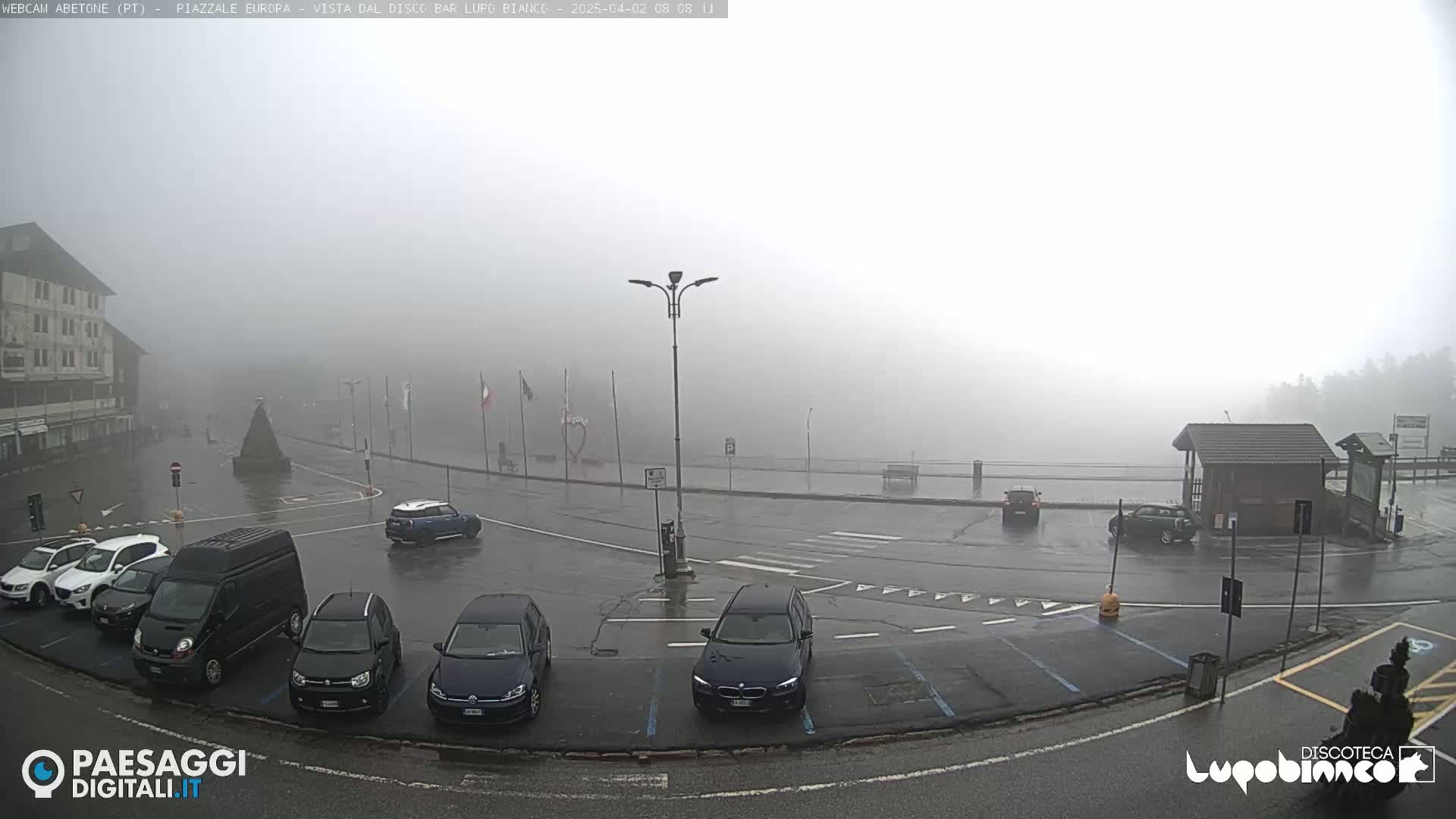 A foggy, rainy parking lot with several cars and a building in the background.