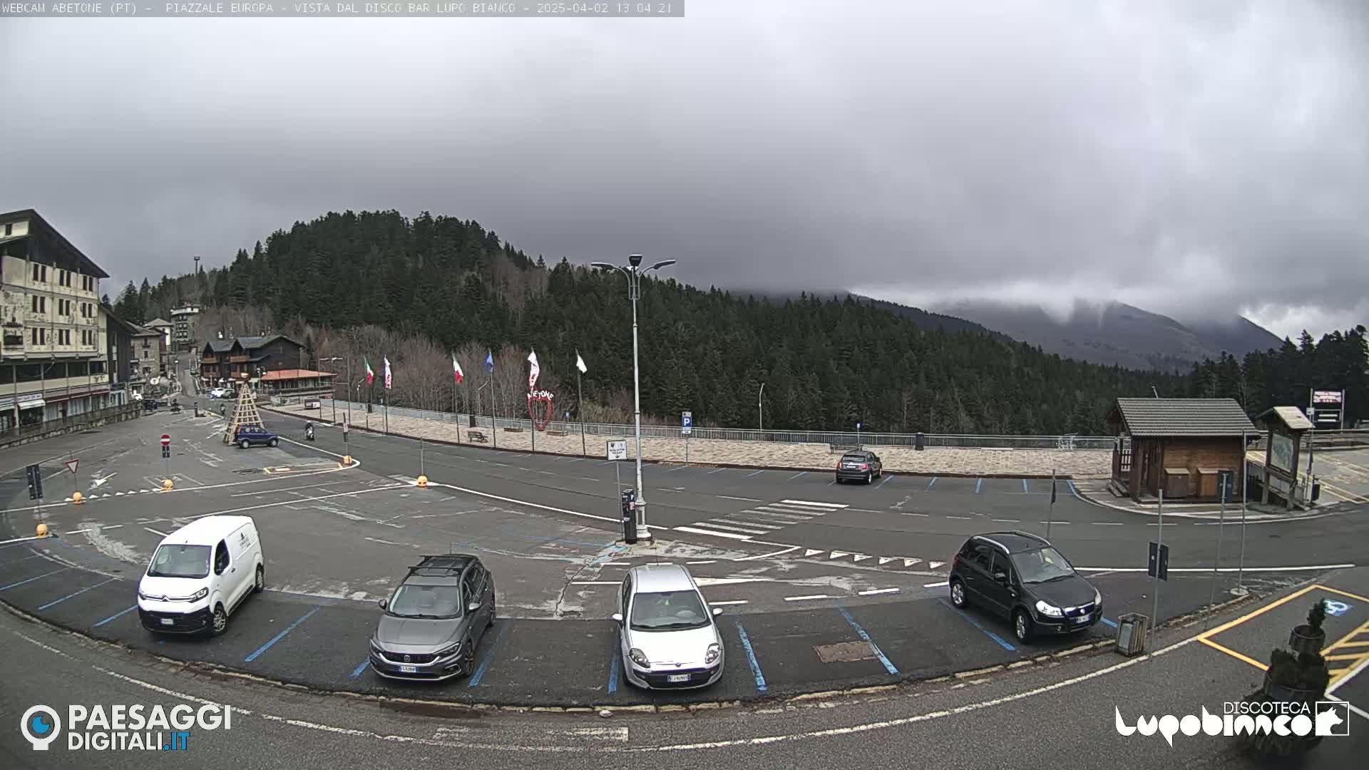 A mostly empty parking lot with several cars, a van, and a small building is situated in a mountain town under a cloudy sky, with a forest and mountain range in the background.