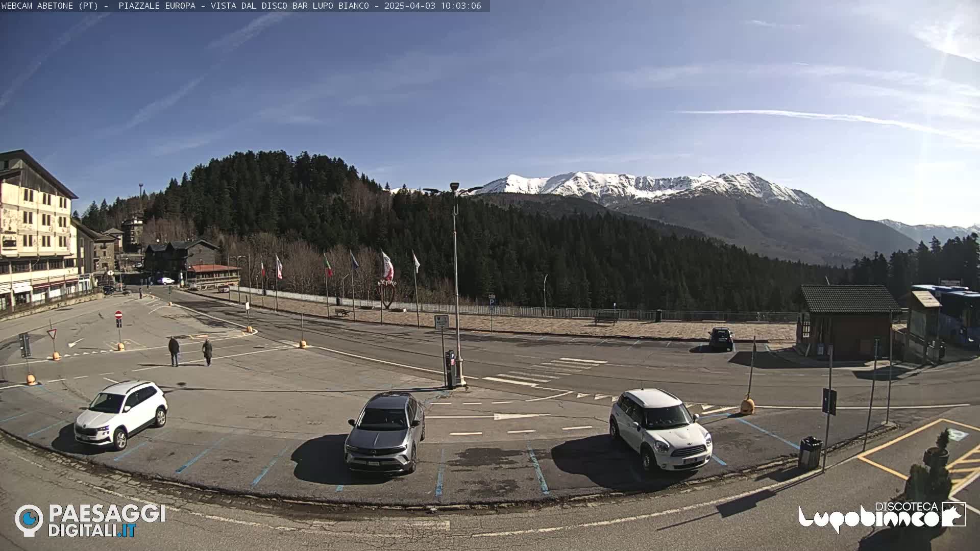 A sunny day in a mountain town parking lot with several cars parked, overlooking snow-capped mountains in the distance.