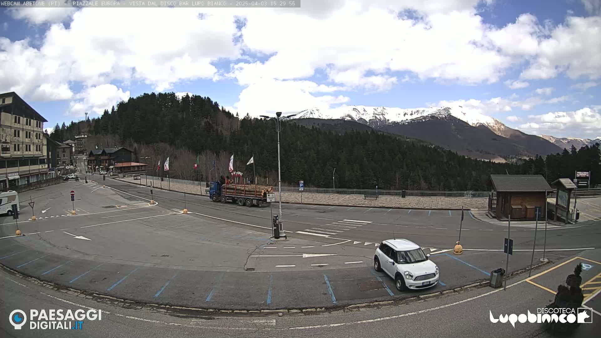 A mostly sunny day in a mountain town parking lot with a few cars and a truck carrying logs, overlooking snow-capped mountains in the distance.