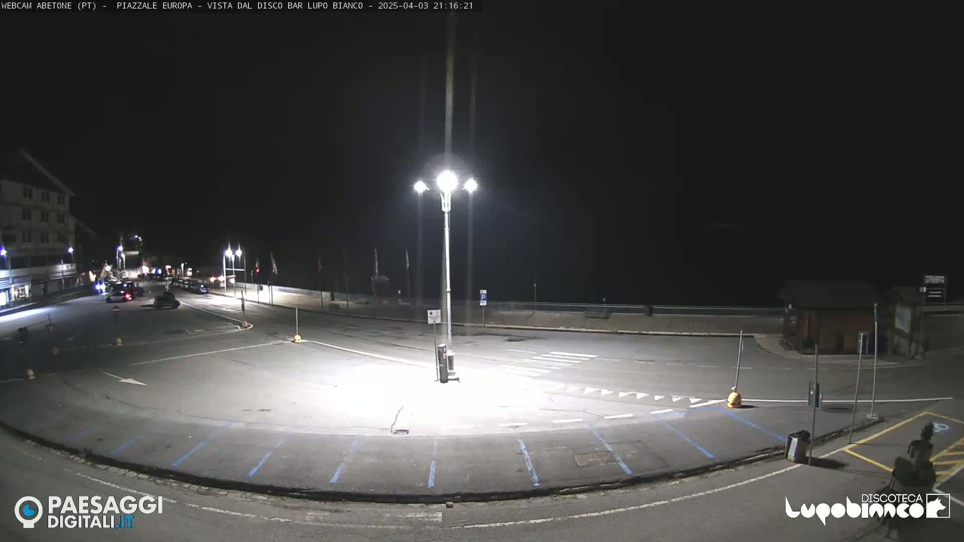An empty, dark parking lot and roadway are illuminated by bright streetlights at night.