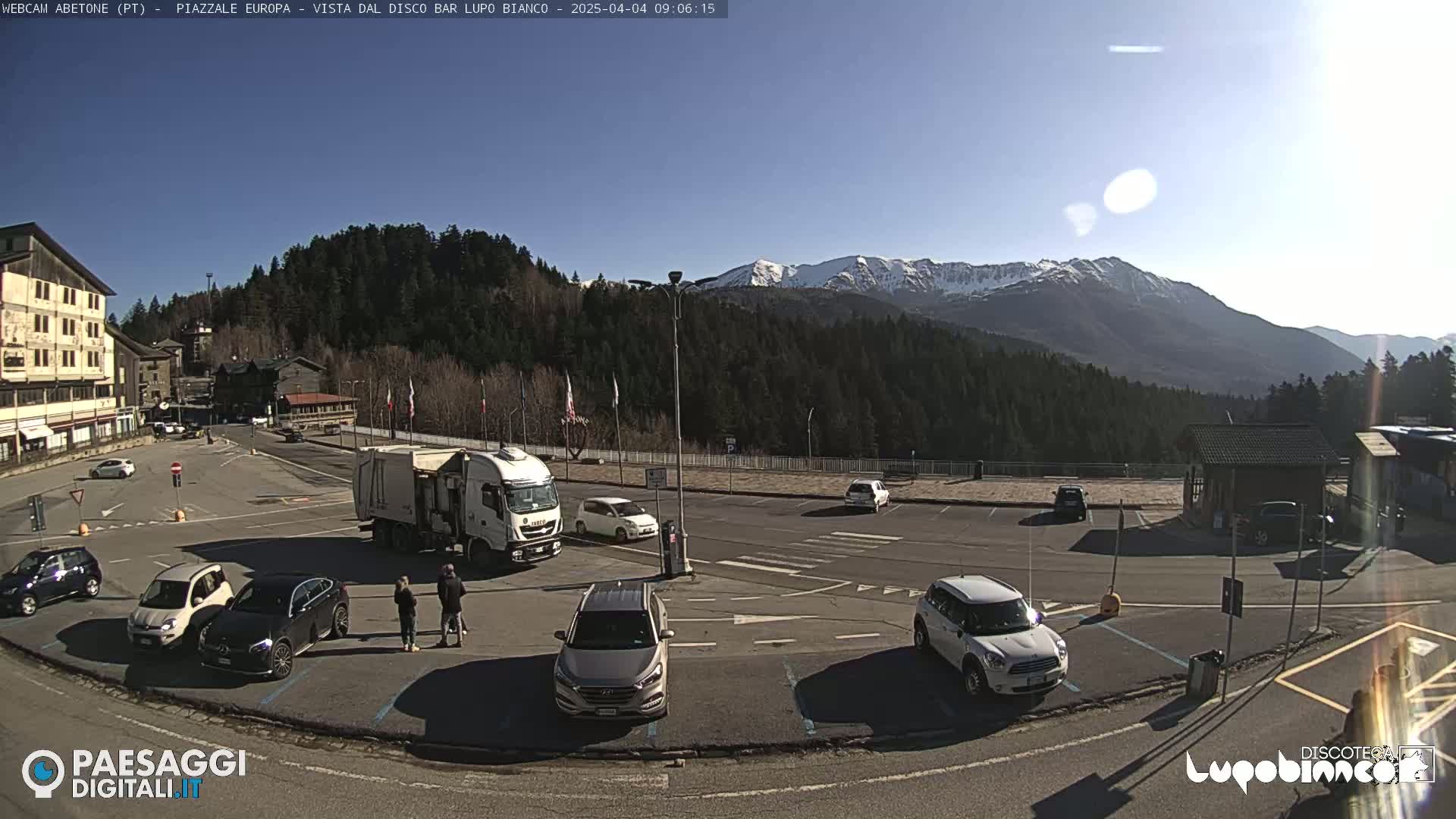 A sunny day in a mountain town parking lot with several cars parked, a large truck, and a few people standing near some of the vehicles, all with snow-capped mountains in the background.