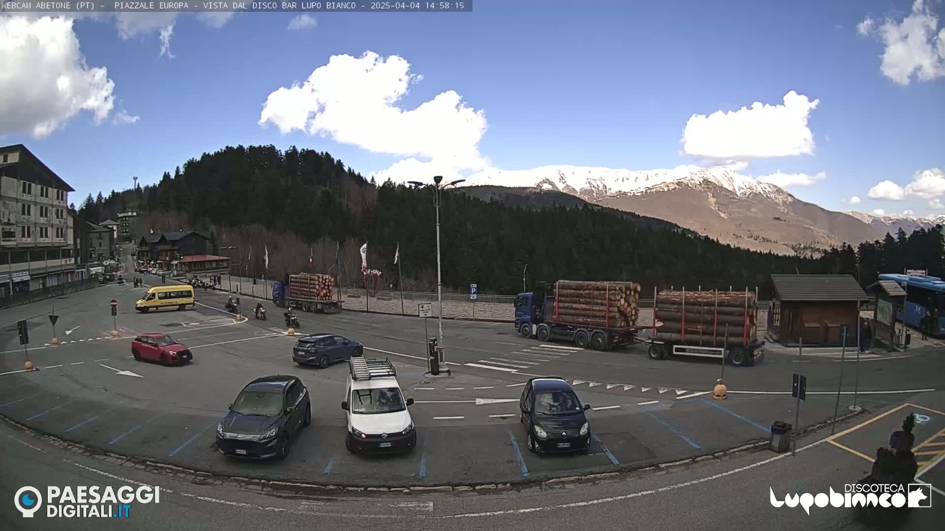 A mostly sunny day shows a parking lot with several cars and trucks carrying logs, near a building and forested mountains with some snow.