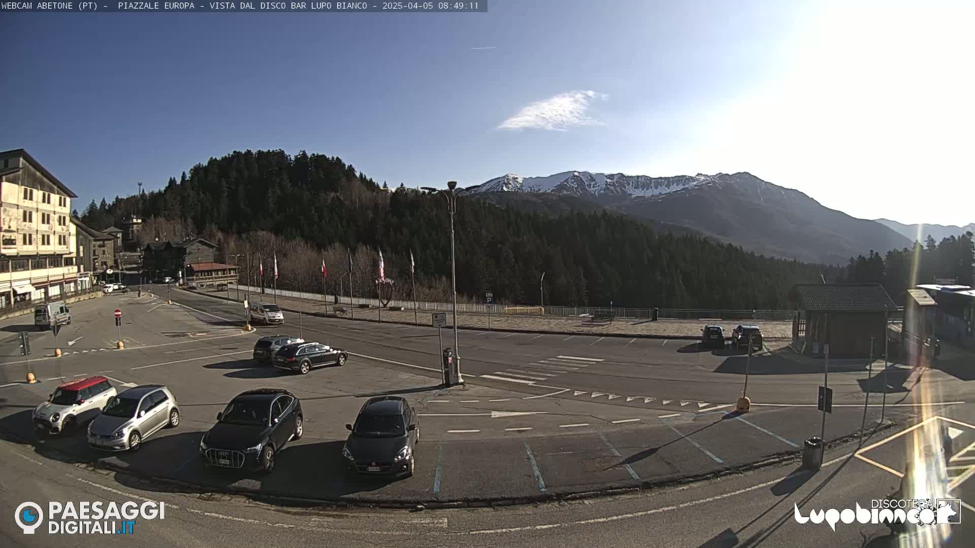 A mostly empty parking lot with several cars is situated in front of buildings and a forested mountain range under a mostly clear, sunny sky.