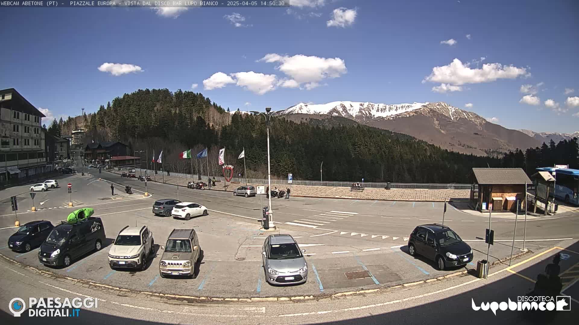 A sunny day reveals a mountain range capped with snow in the background, overlooking a mostly empty parking lot with several cars and a few people near a building.