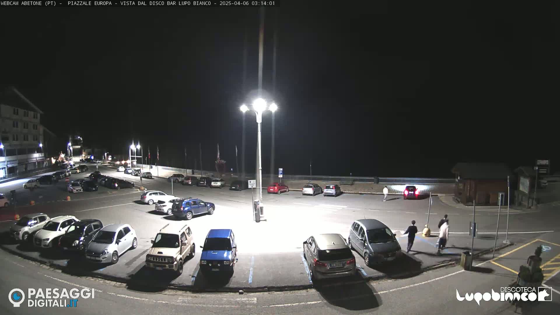 A nighttime view of a mostly empty parking lot with several cars, a few people walking, and a building in the background under bright artificial lights.
