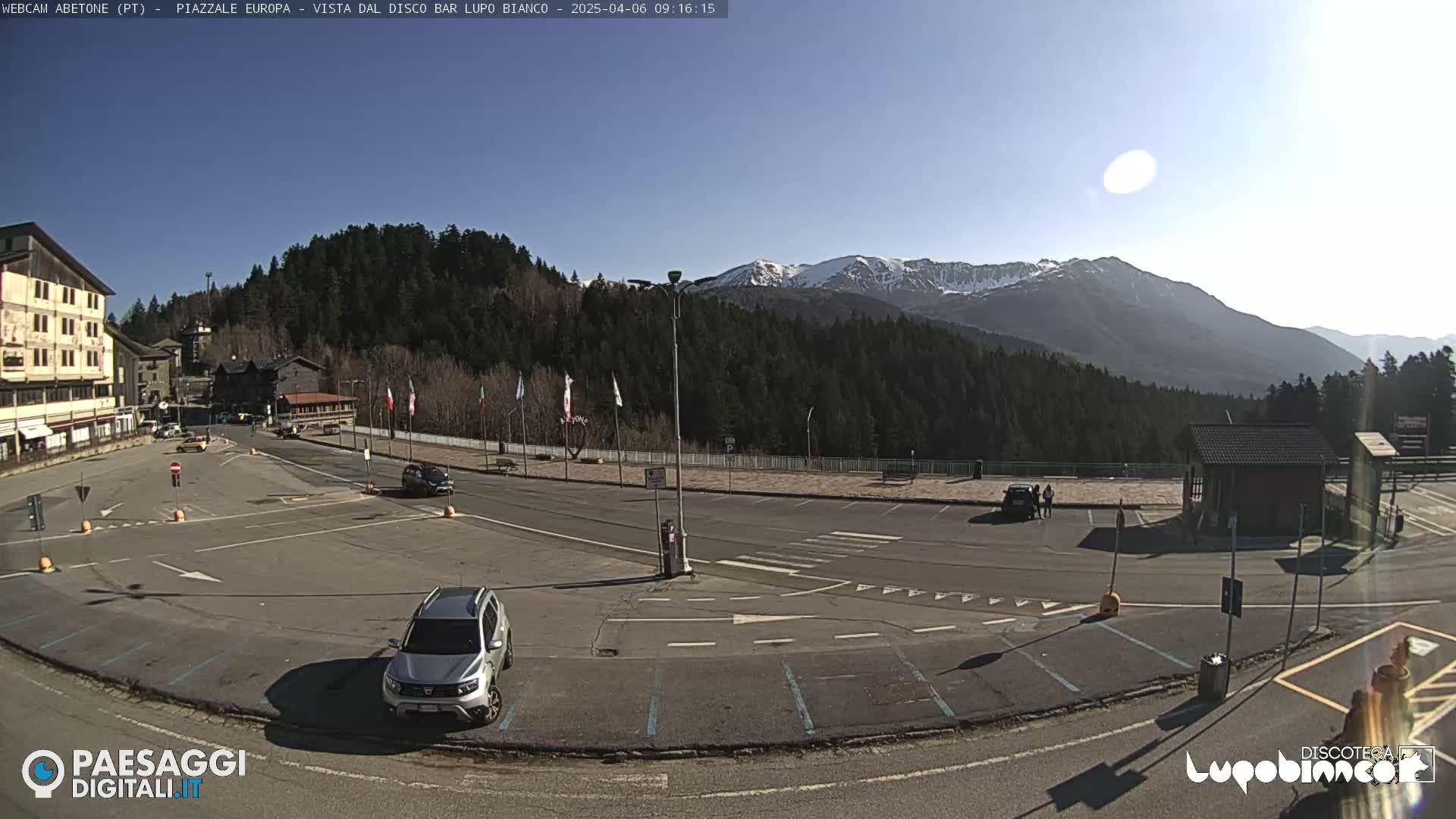 A mostly empty parking lot overlooks a mountain range under a clear, sunny sky.