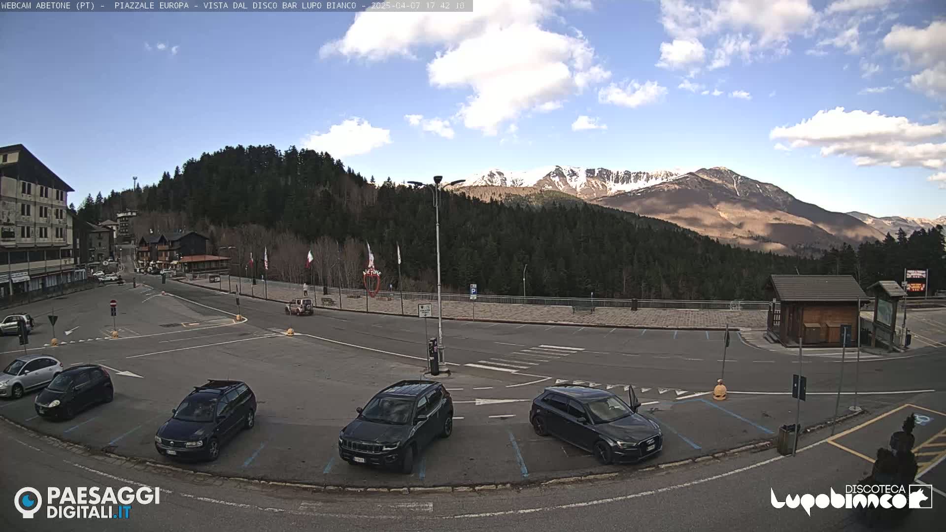 A mostly sunny day shows a parking lot in a mountain town, with several cars parked and a mountain range in the background.