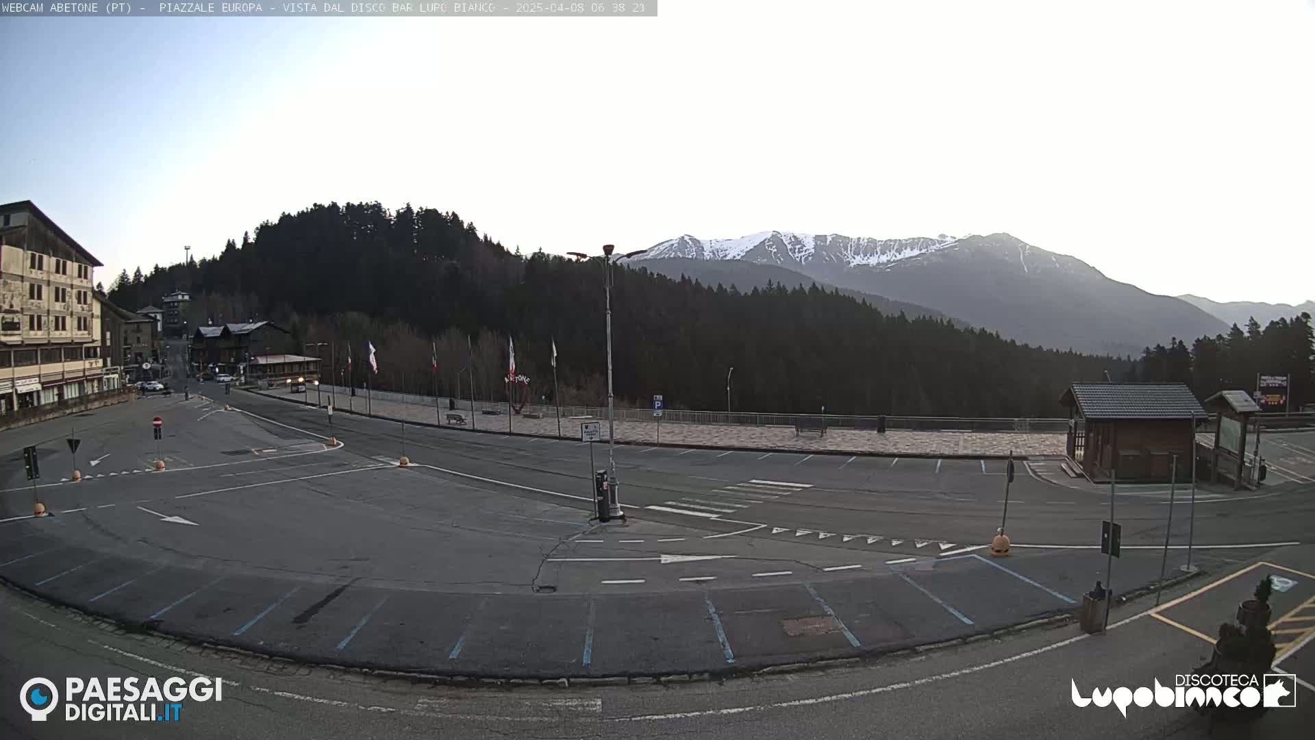 A mostly empty parking lot and plaza overlook a snow-capped mountain range under a clear, bright sky.