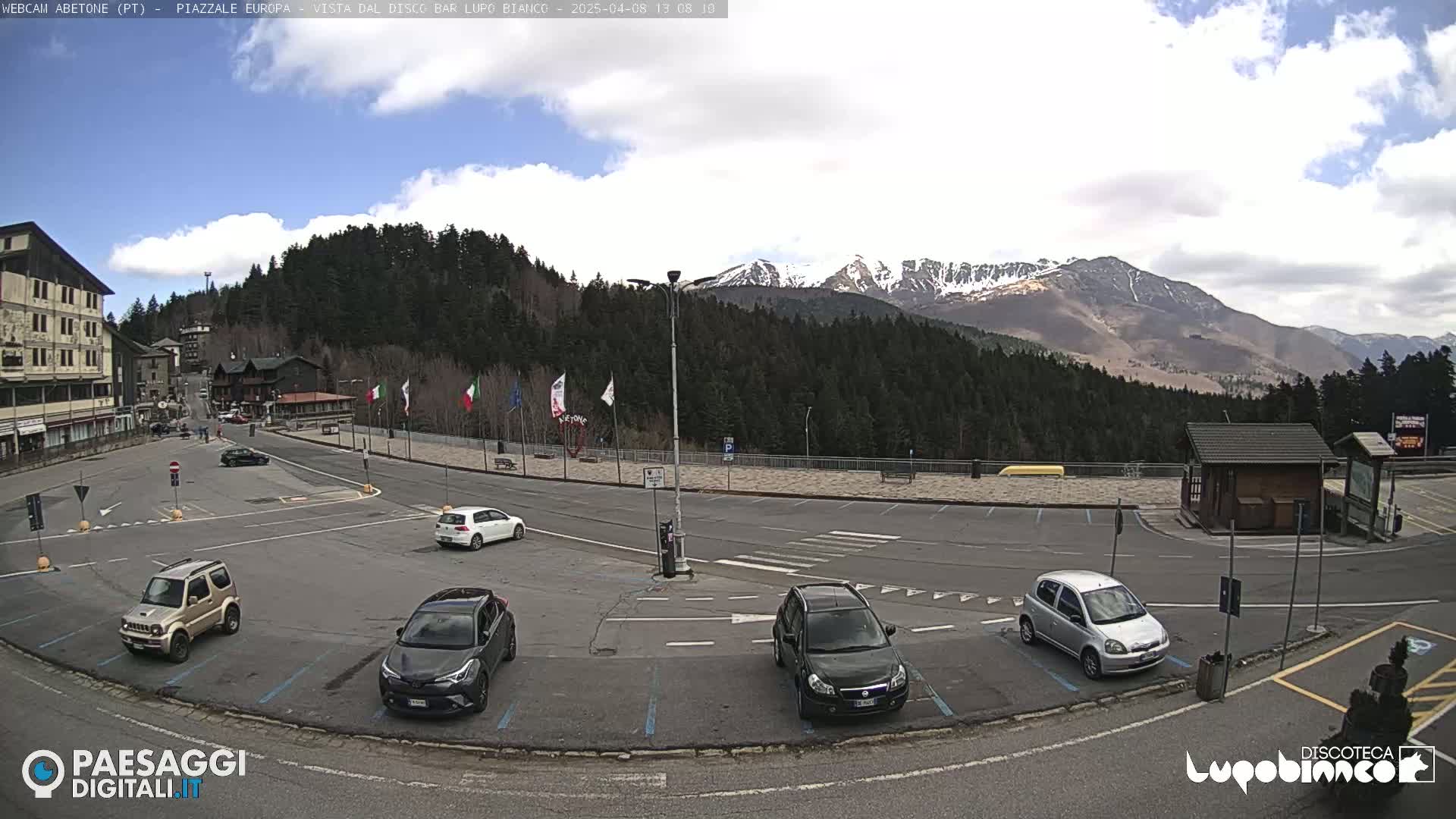 A mostly sunny parking lot with several cars, overlooked by a building and a mountain range with some snow visible in the background.