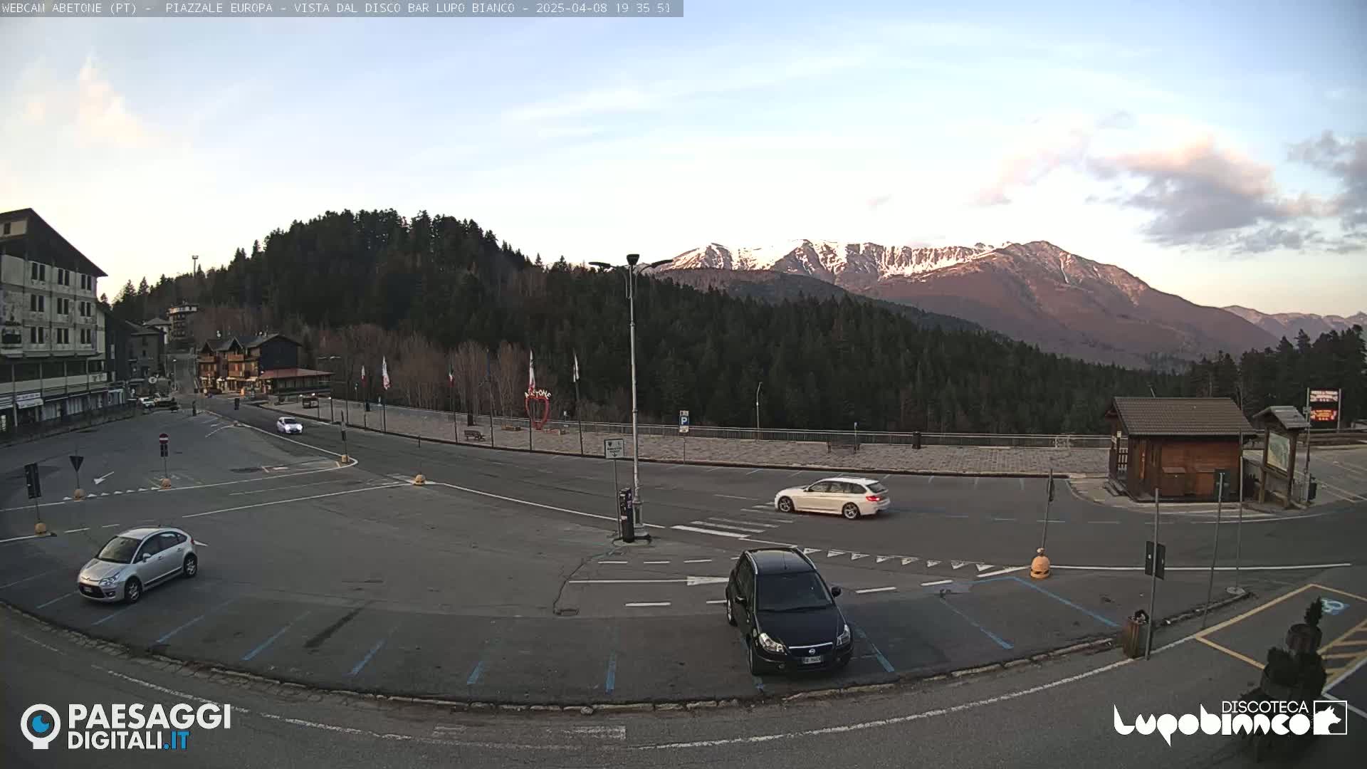 A mostly empty parking lot with a few cars, situated in front of buildings and a mountain range with snow-capped peaks under a partly cloudy sky.