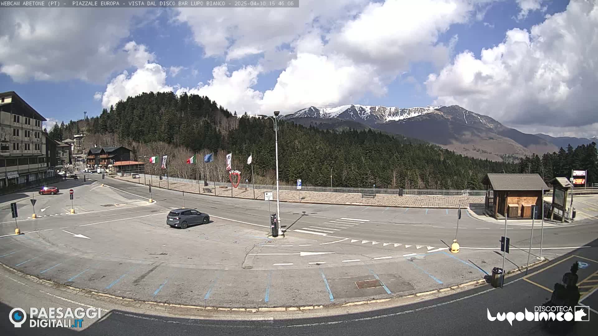 A mostly empty parking lot with a few cars is situated near buildings and a roadway, overlooking a mountain range with patches of snow under a partly cloudy sky.