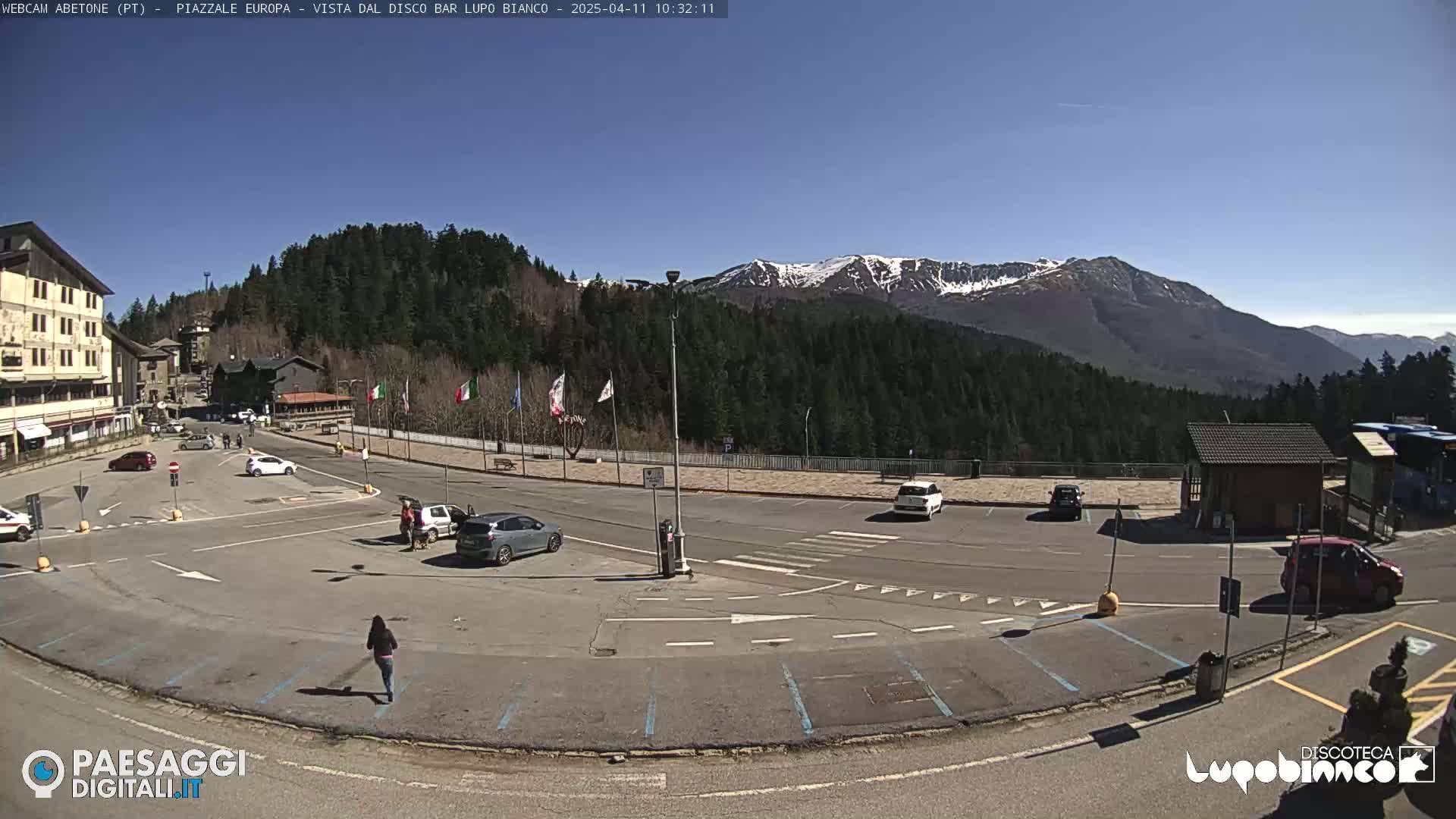 A mostly empty parking lot with several cars and a few people is situated in front of buildings and trees, overlooking snow-capped mountains under a clear blue sky.