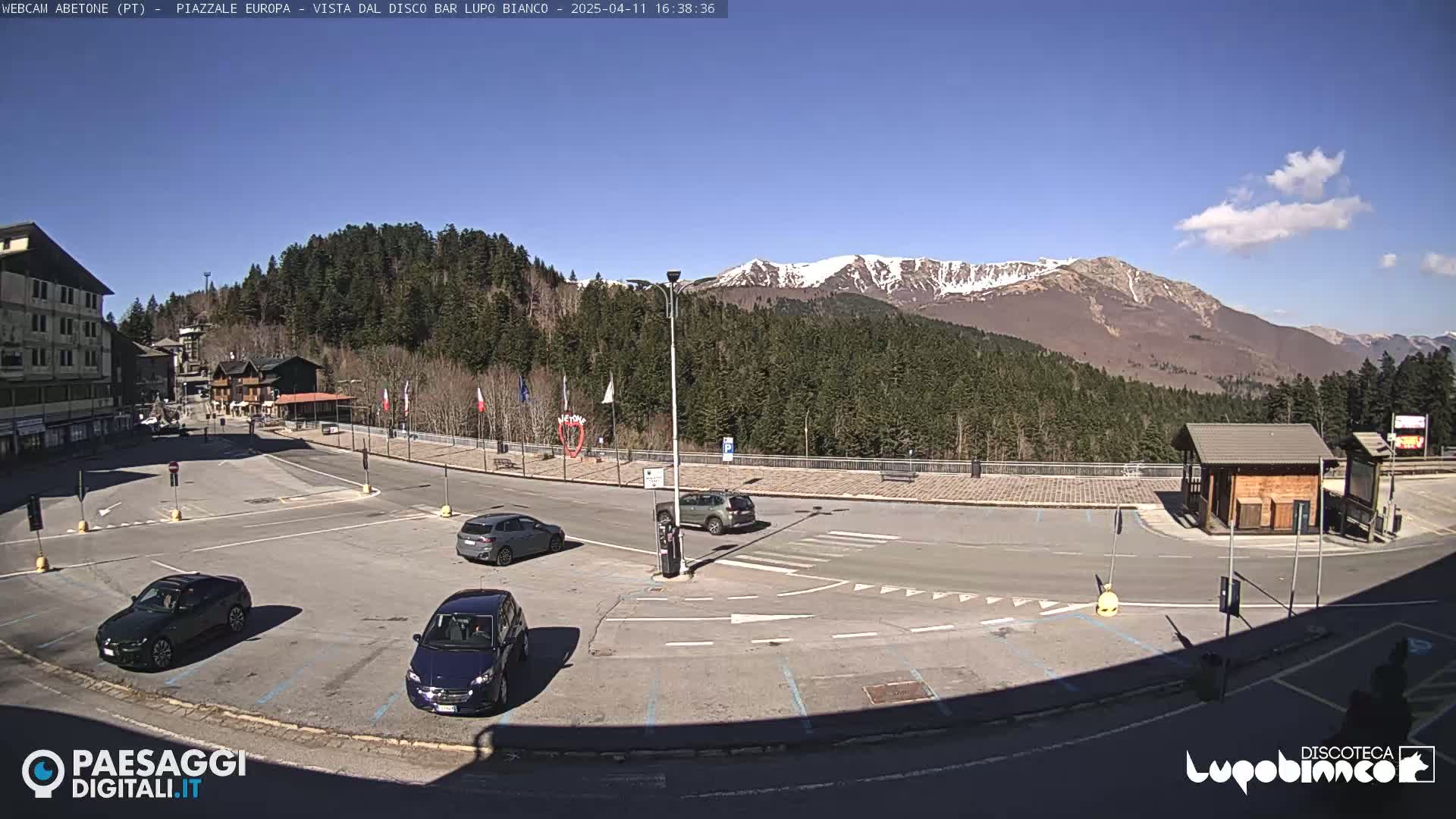 A mostly sunny parking lot with several cars, overlooked by buildings and a snow-capped mountain range in the background.