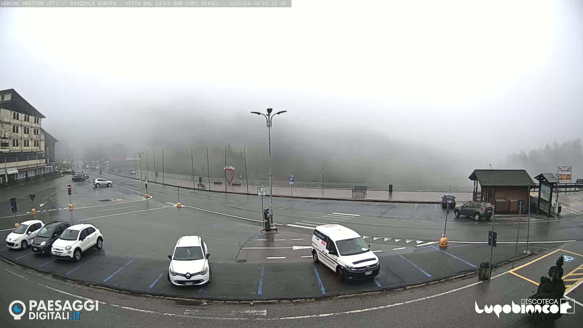 A wet, foggy parking lot with several cars and a van is surrounded by a building and a hill covered in fog.