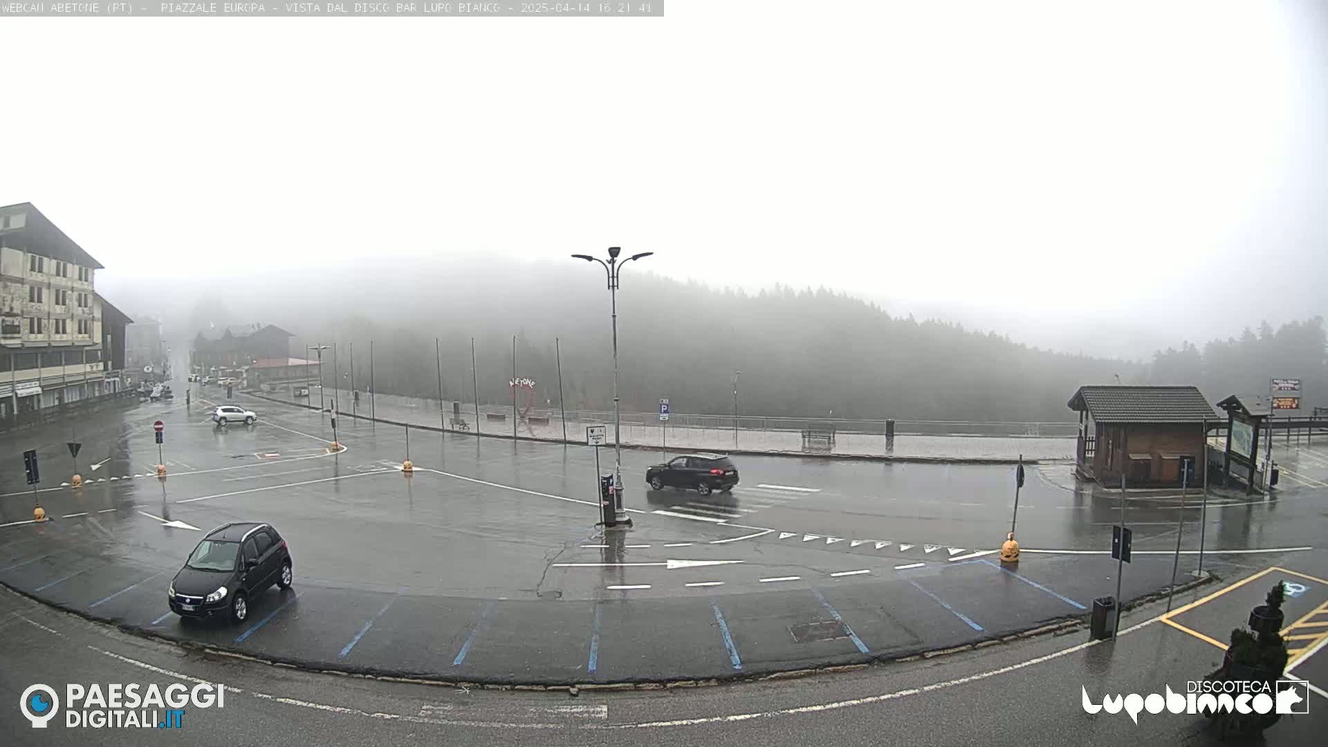 A wet, foggy parking lot with several cars and a building in the background.