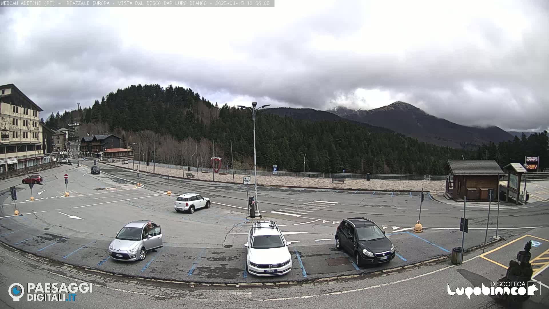 A mostly empty parking lot with several cars, situated in front of buildings and a forest with mountains in the background under an overcast sky.