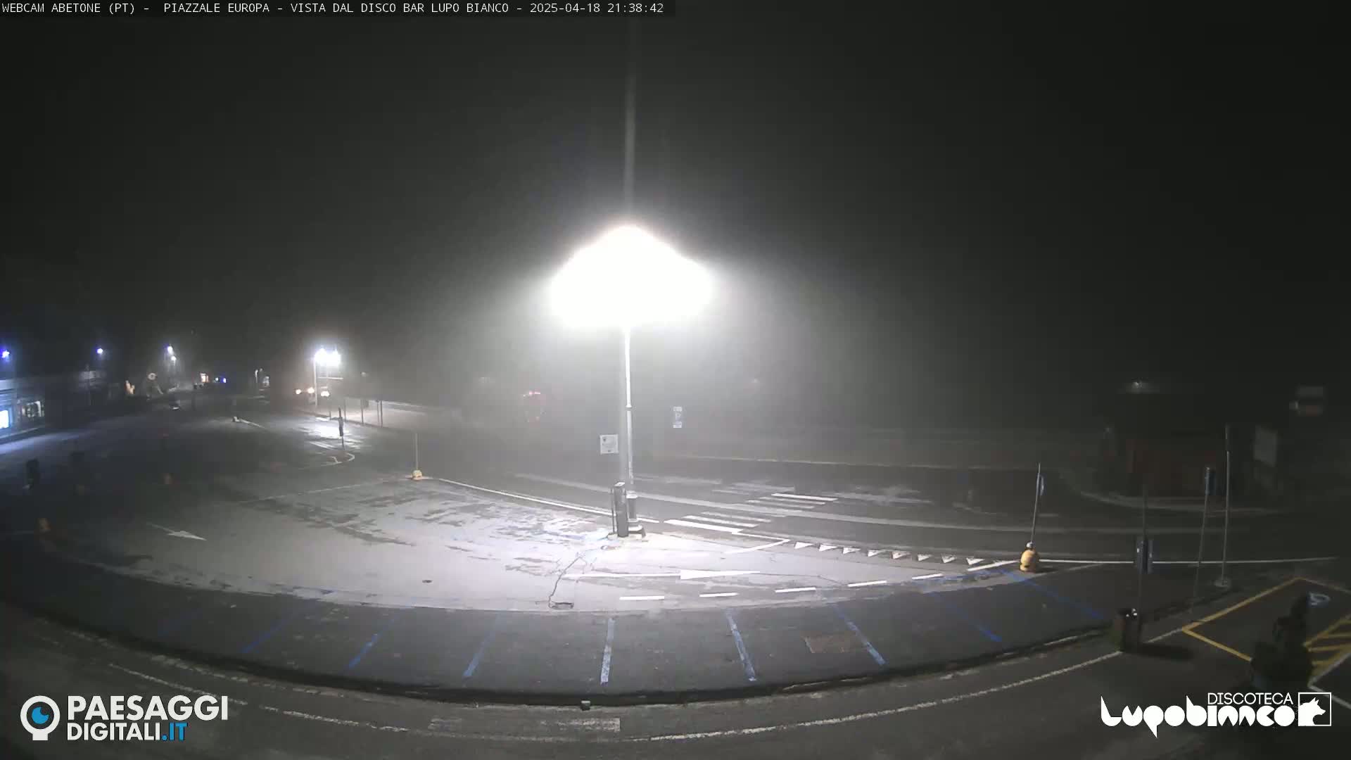 A foggy, nighttime view of an empty parking lot and roadway intersection illuminated by bright streetlights.