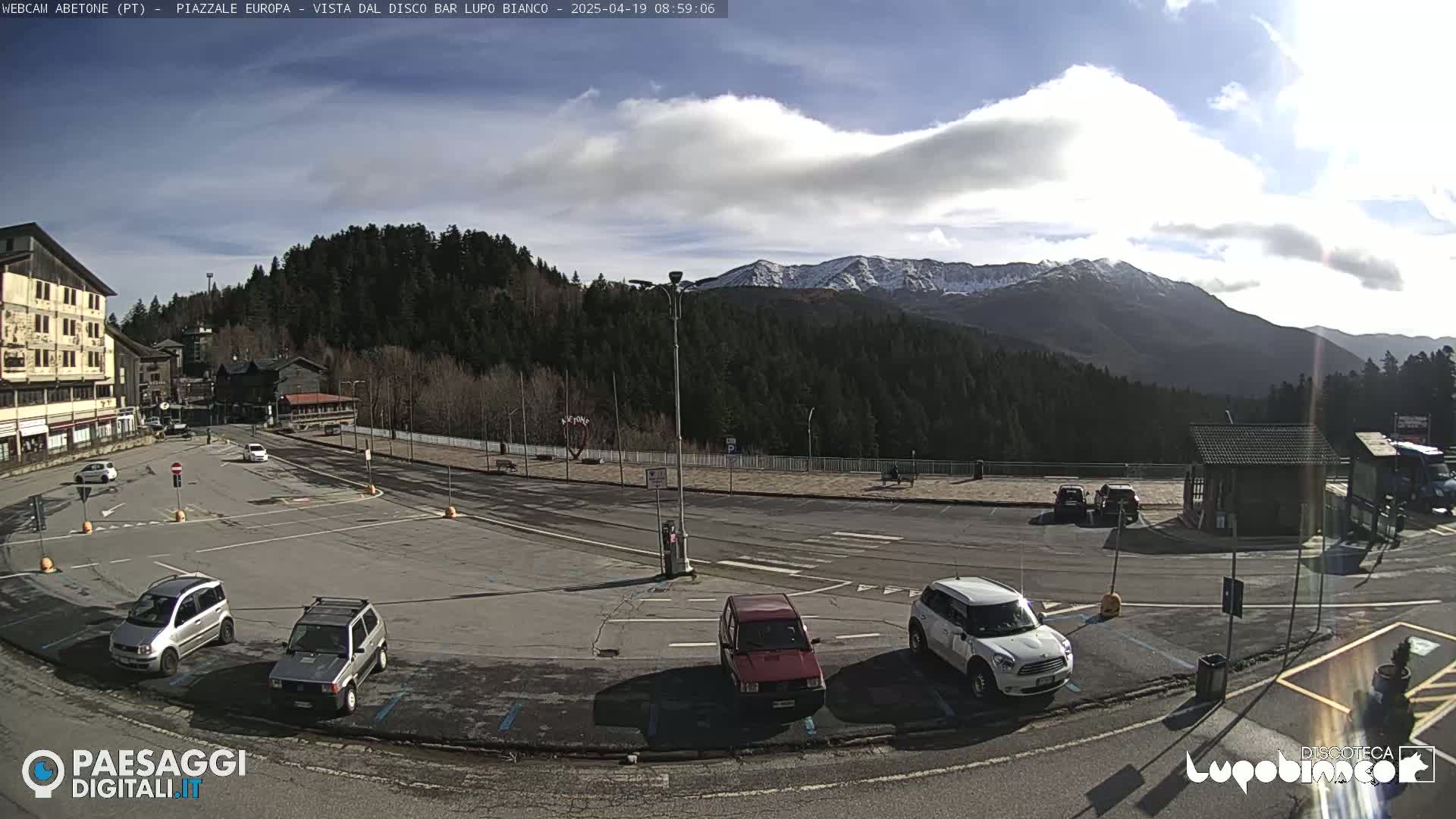 A mostly empty parking lot with several cars is situated in front of a building, overlooking a snow-capped mountain range under a partly cloudy, sunny sky.