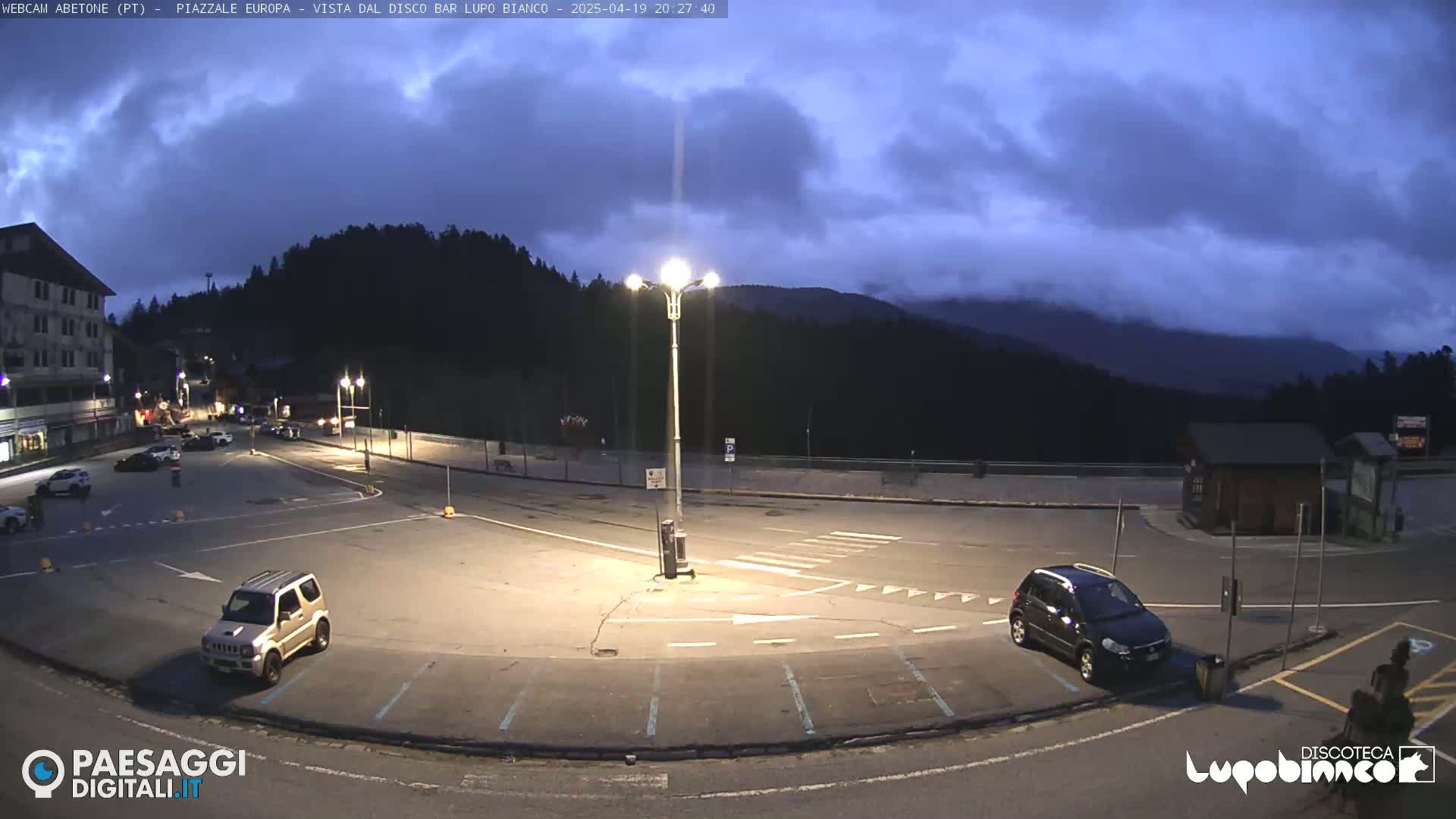 An empty parking lot at dusk, with a few cars parked, is situated near a building and overlooks a mountain range under a cloudy sky.