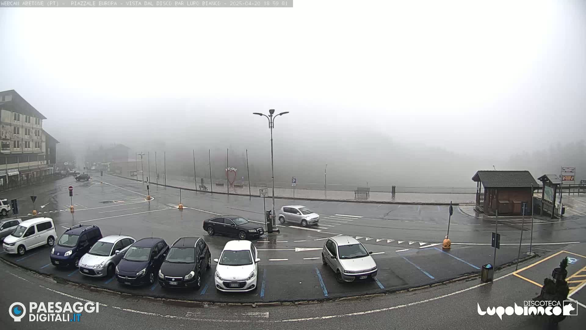 A foggy parking lot with several cars parked in it, next to a building and a small wooden structure, all wet from rain.