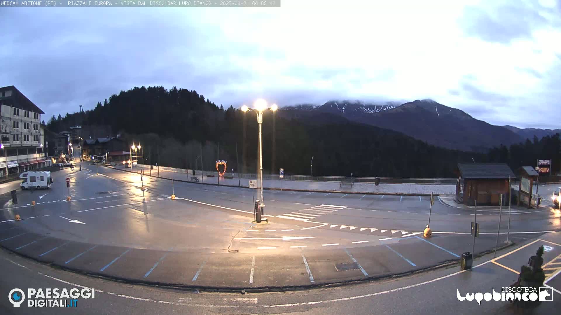 An empty, wet parking lot is situated in front of a mountain range under an overcast sky.