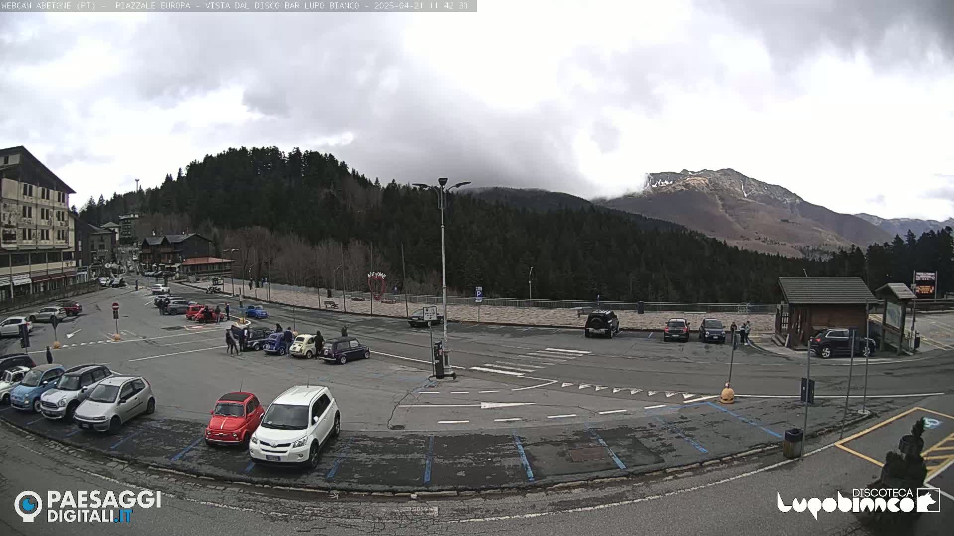 A parking lot with several cars is situated in front of a building and overlooks a mountain range partially covered in clouds and snow under an overcast sky.