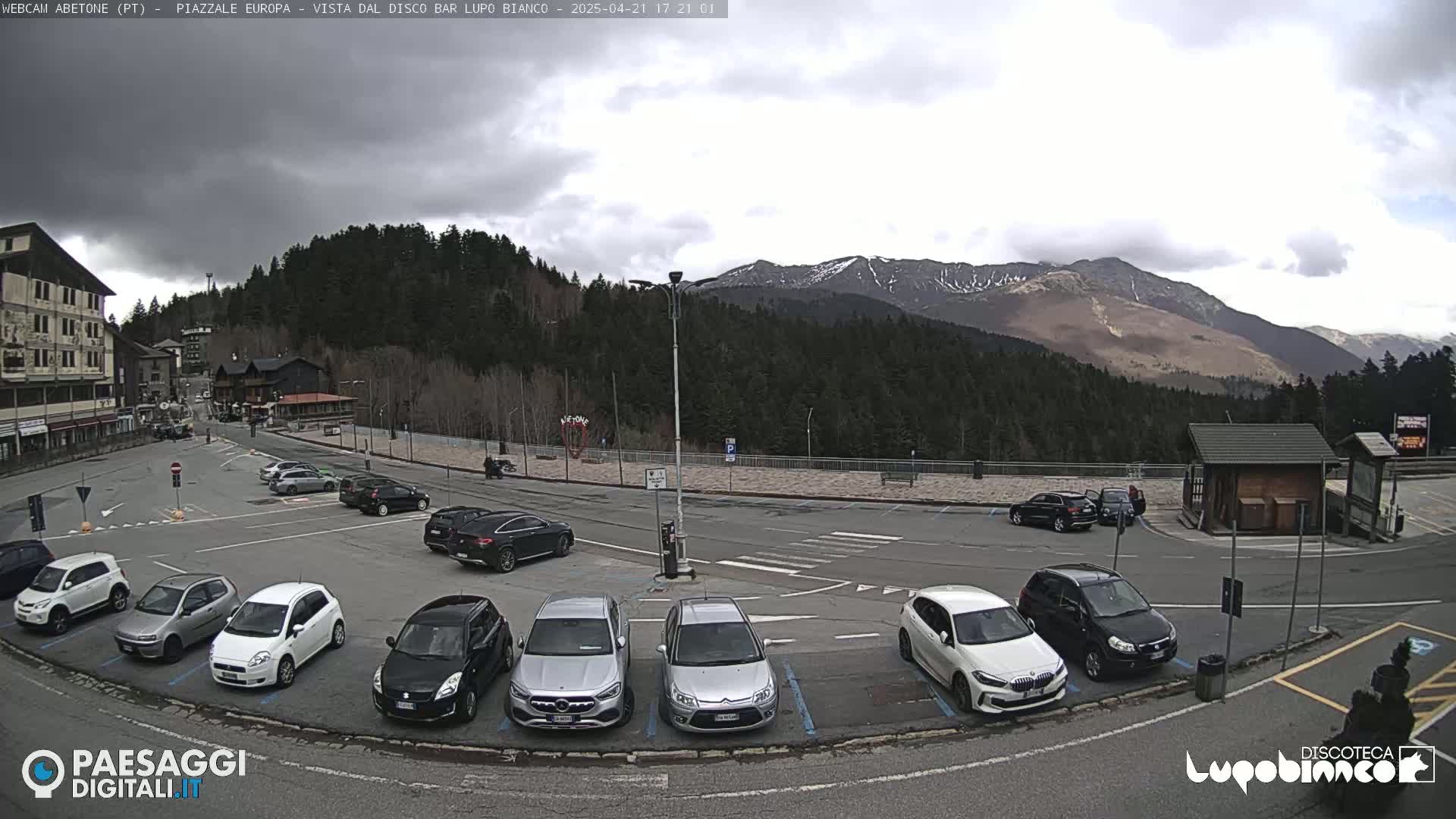 A mostly cloudy day reveals a parking lot full of cars in front of a mountain range partially covered in snow.