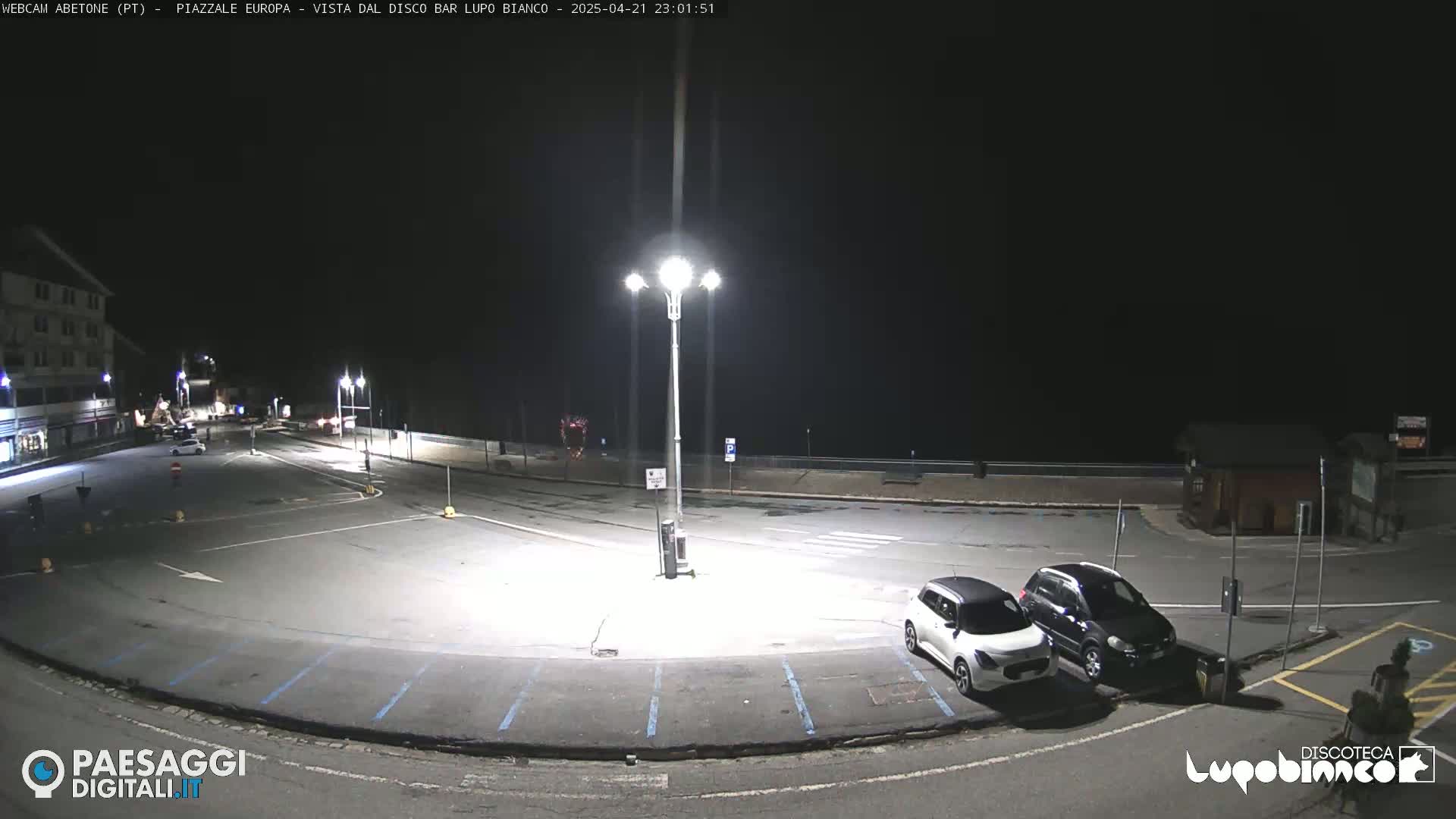 A nighttime view of an empty parking lot with a few cars, illuminated by streetlights, next to a building.