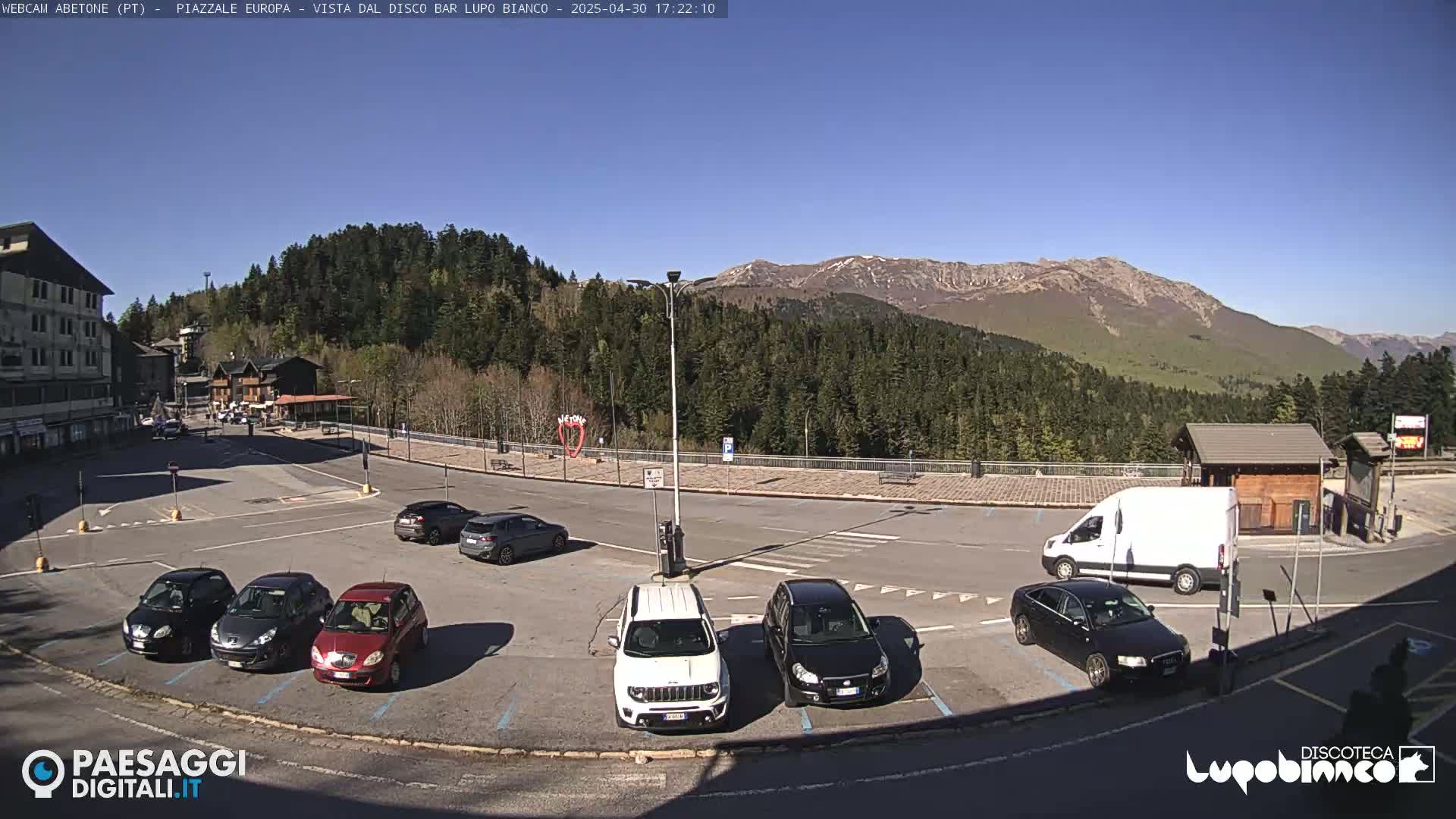 A mostly empty parking lot with several cars and a van is situated in front of a building, overlooking a mountain range under a clear blue sky.