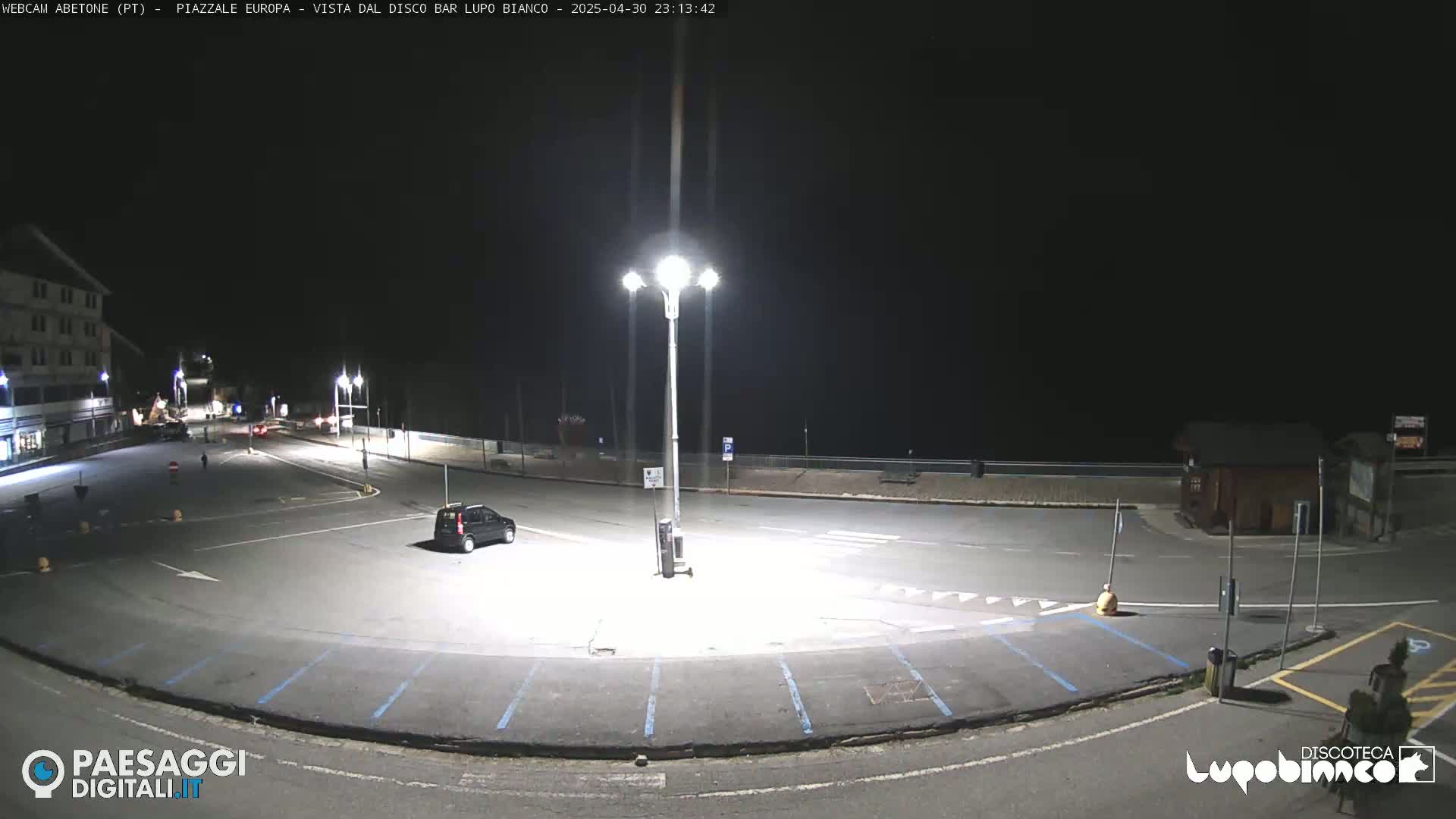 A mostly empty nighttime parking lot is illuminated by a tall lamppost, with a few cars visible and a building in the background.