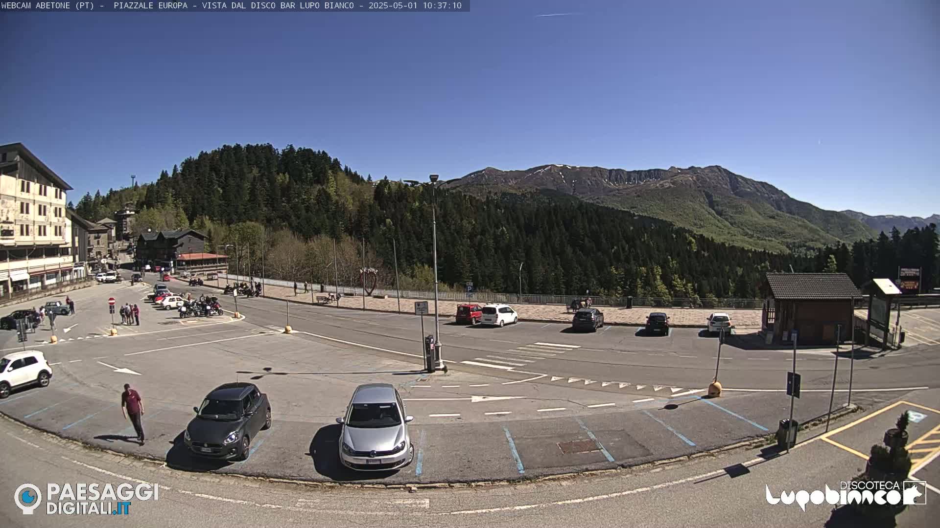 A sunny day in a mountain parking lot with several cars, a few pedestrians, and a background of green hills and mountains.