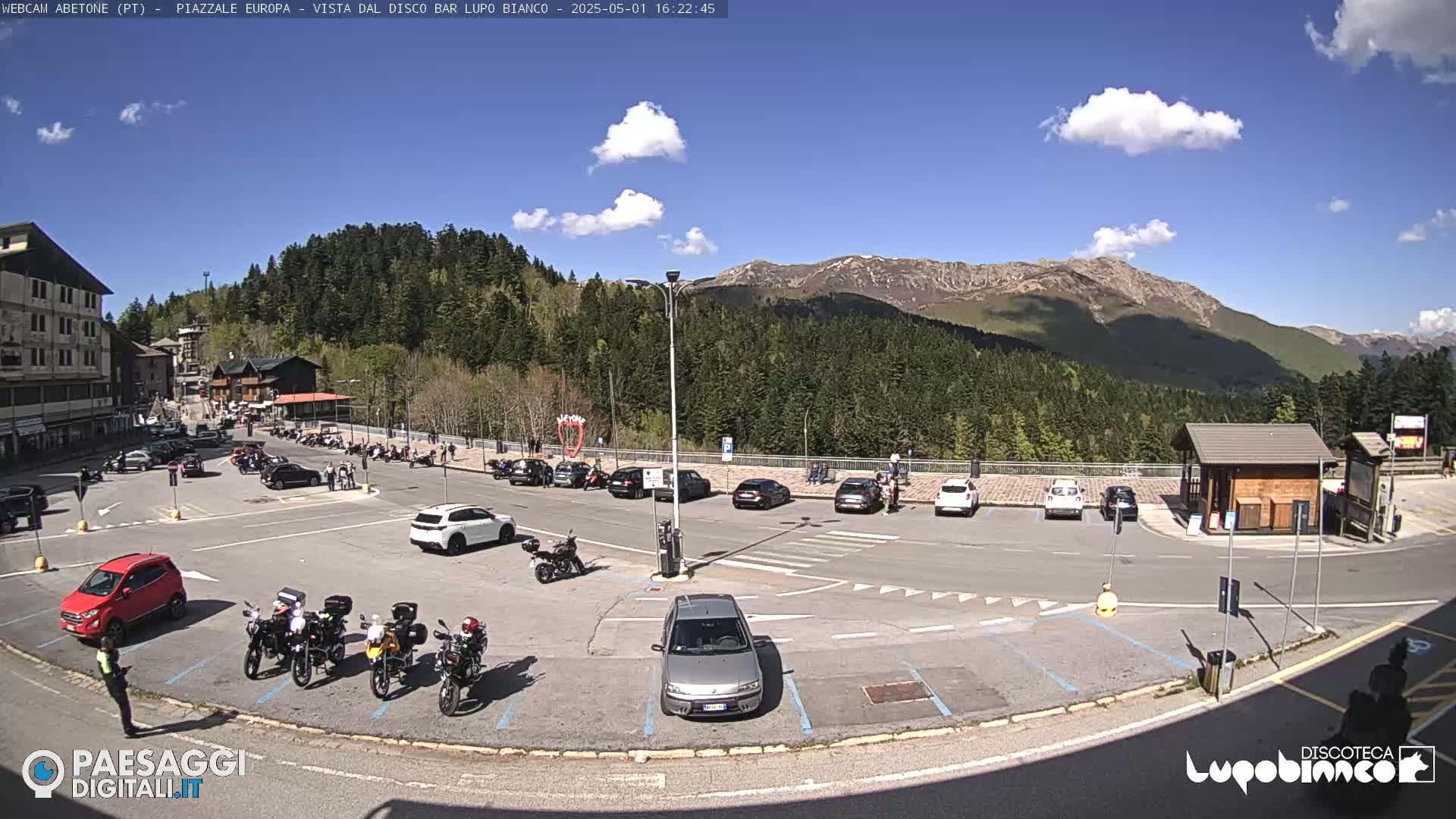 A sunny day in a mountain parking lot with several cars and motorcycles parked, a few people walking around, and a mountain range in the background.