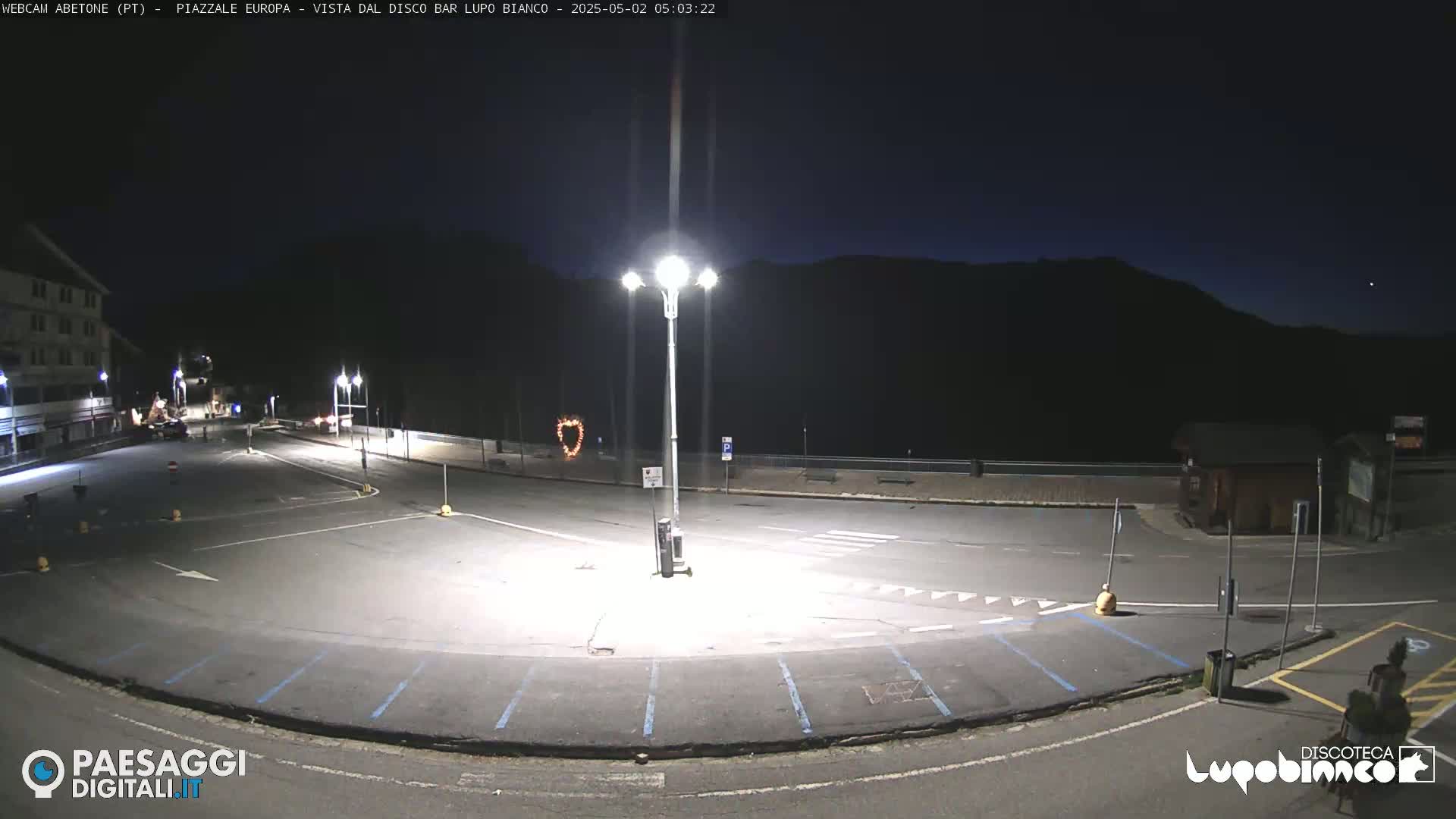 A mostly empty, dark parking lot is illuminated by bright streetlights at night, with a building and a mountain range in the background.