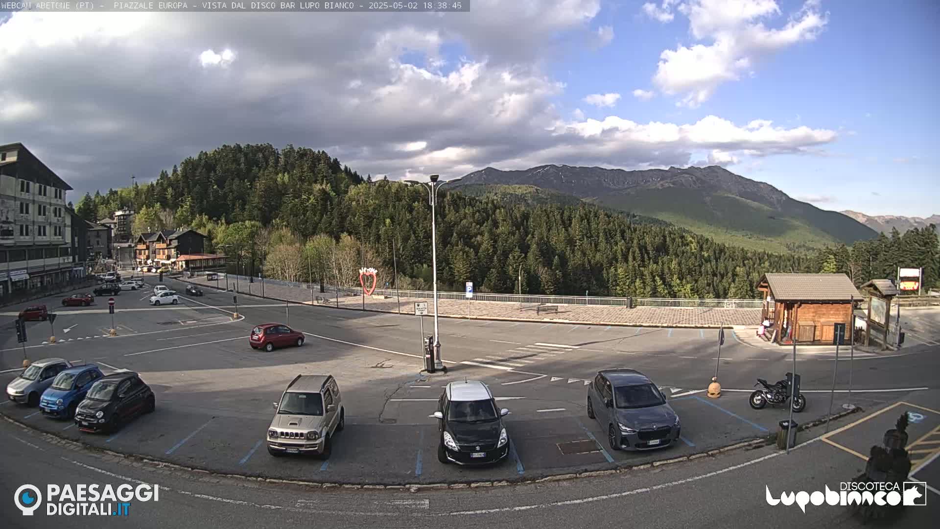 A mostly empty parking lot with several cars is situated in front of a building and a small wooden structure, overlooking a mountain range under a partly cloudy sky.