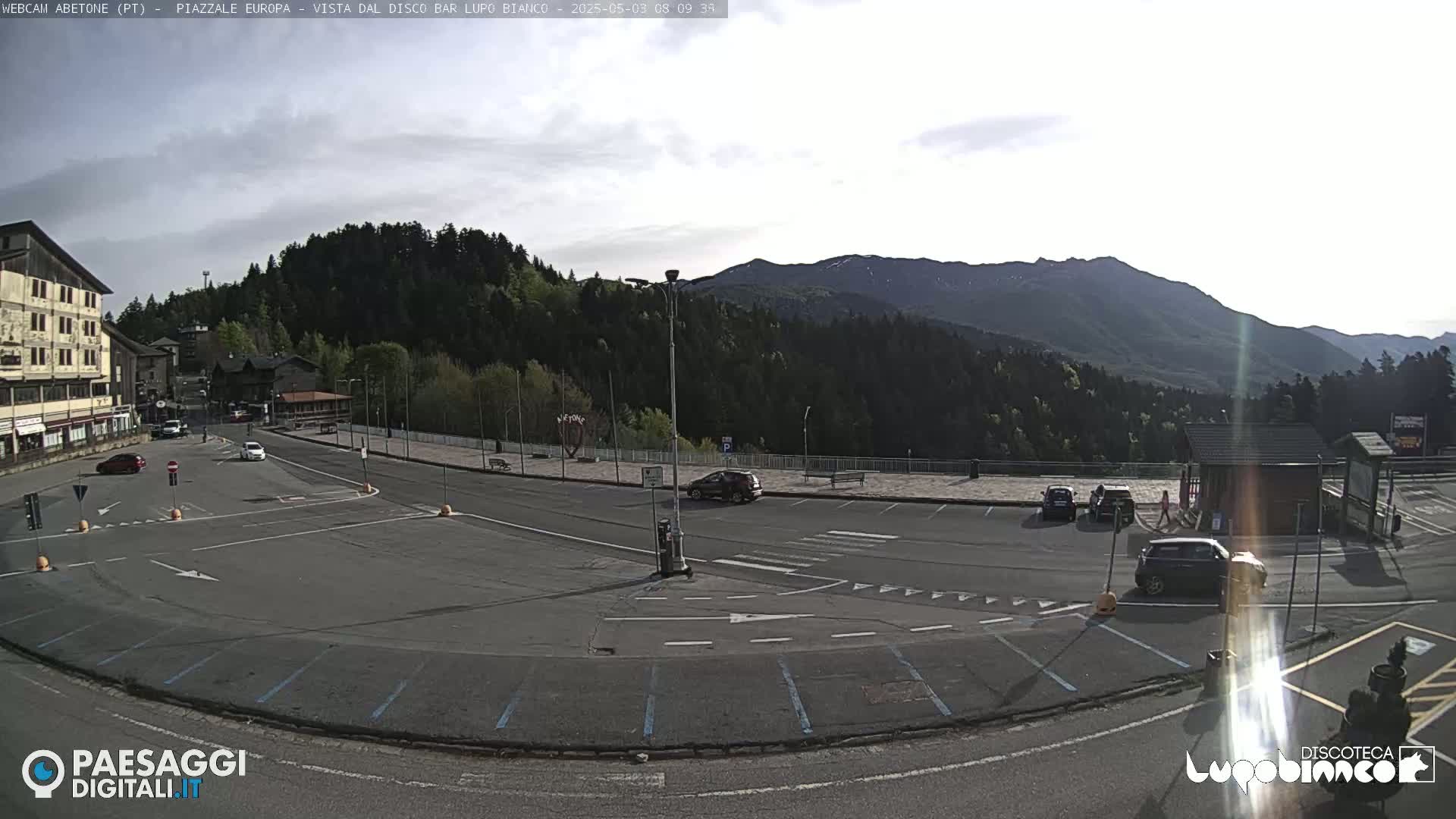 A mostly empty parking lot is situated in front of buildings and forested mountains under a partly cloudy sky.