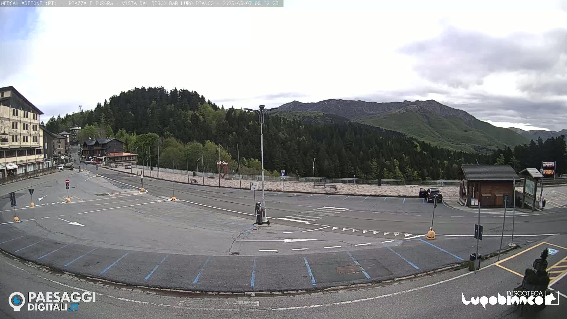 An empty parking lot with light gray pavement and blue parking lines is situated in front of buildings and a forested mountain range under a cloudy sky.