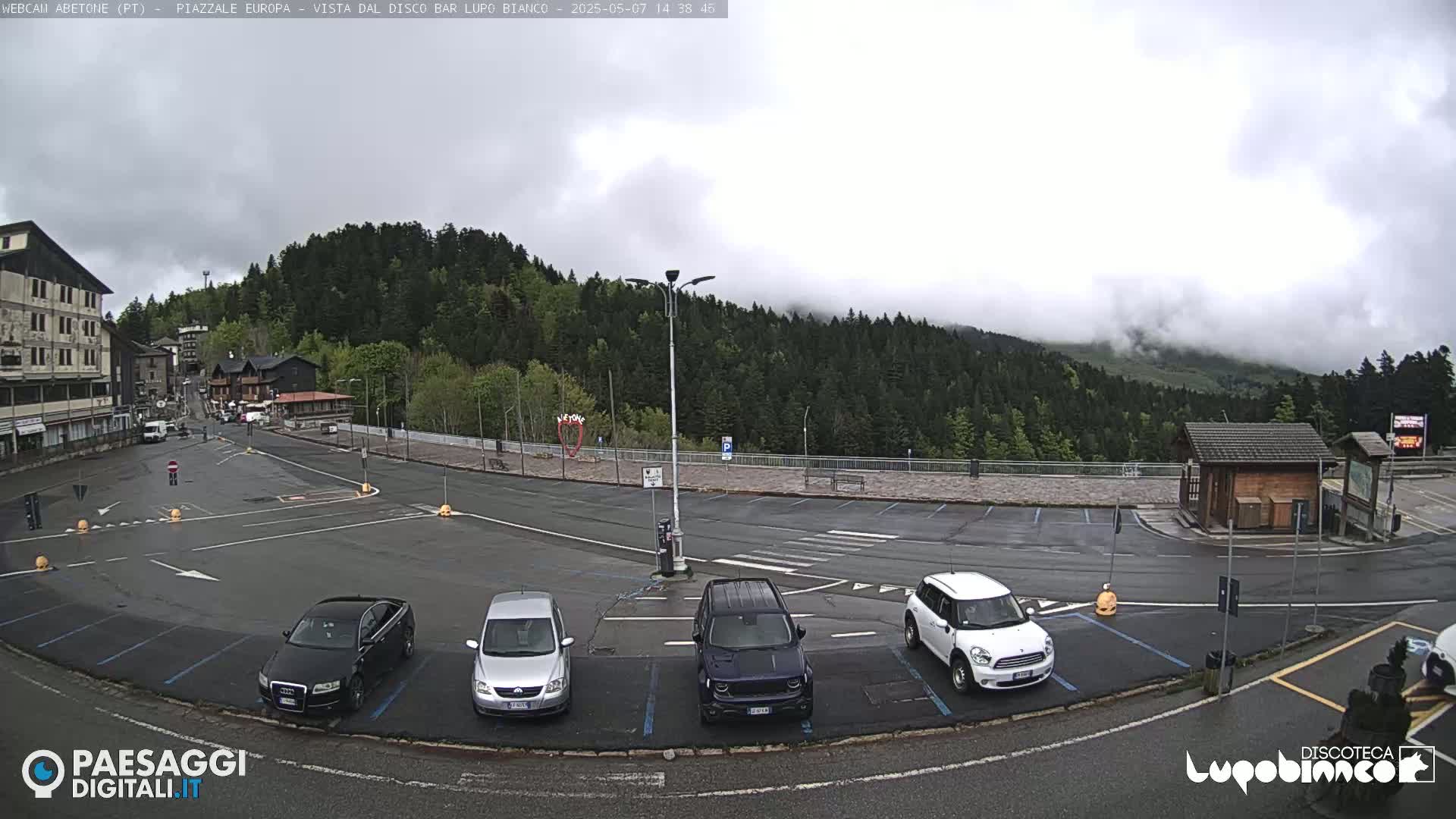 A mostly empty parking lot with several cars is situated in front of a building and overlooks a mountain range partially obscured by fog.