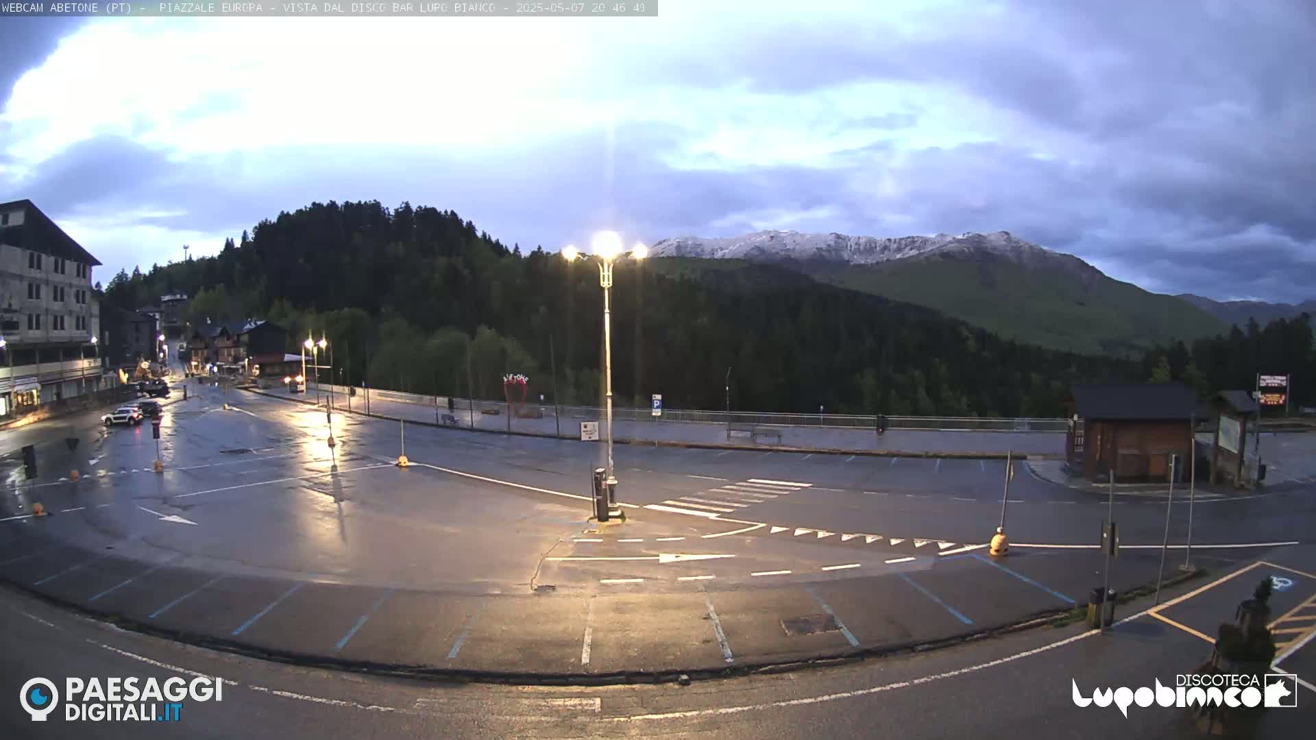 A wet, empty parking lot and roadway are situated in a mountain valley under a partly cloudy, twilight sky; snow is visible on distant peaks.