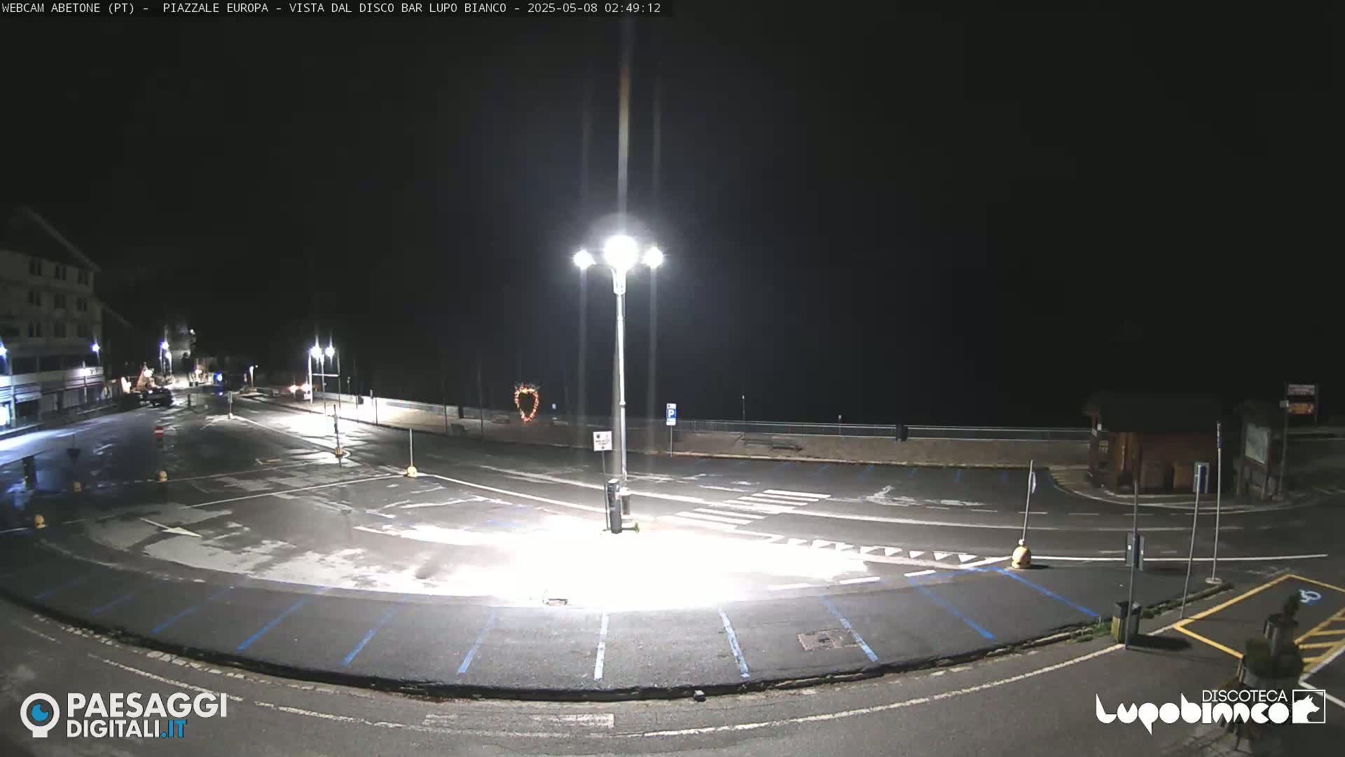 An empty, dark parking lot at night, illuminated by streetlights, is wet from recent rain or moisture.
