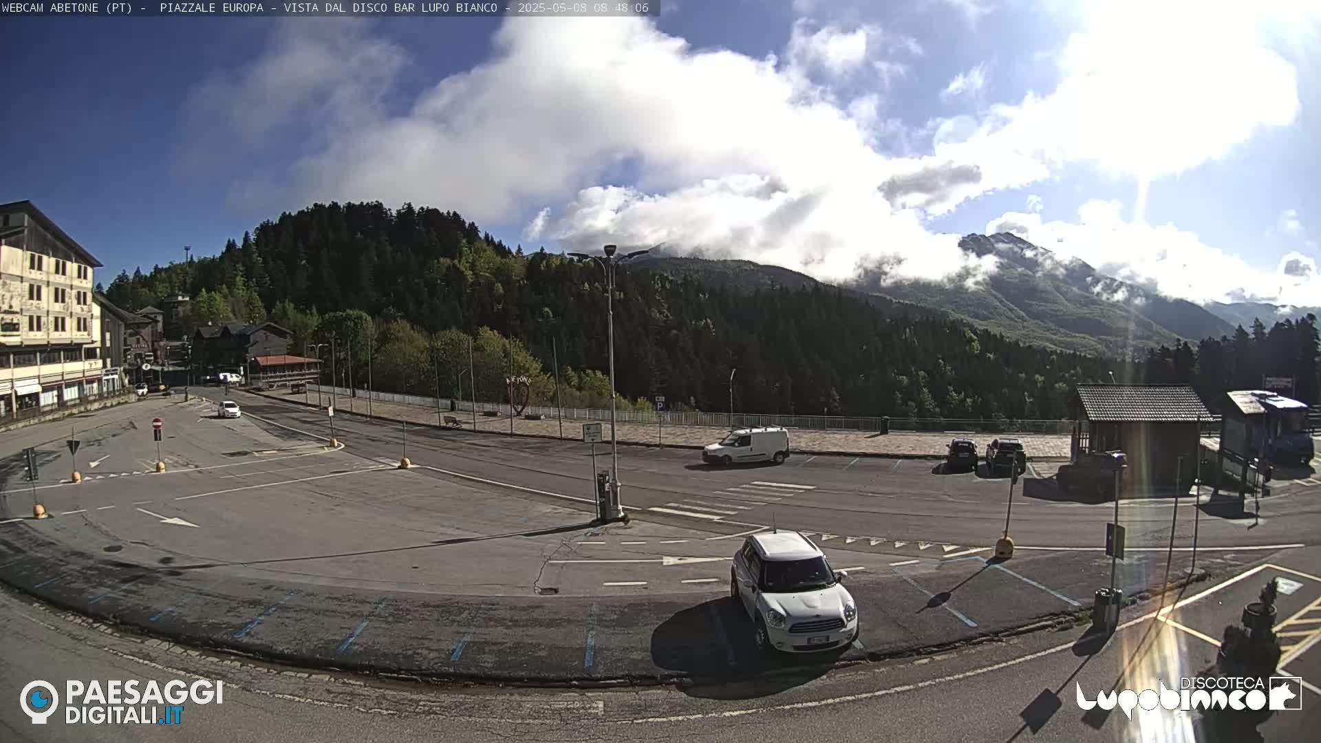 A mostly sunny parking lot overlooks a mountain range partially obscured by clouds, with several cars parked and a few buildings visible in the foreground.