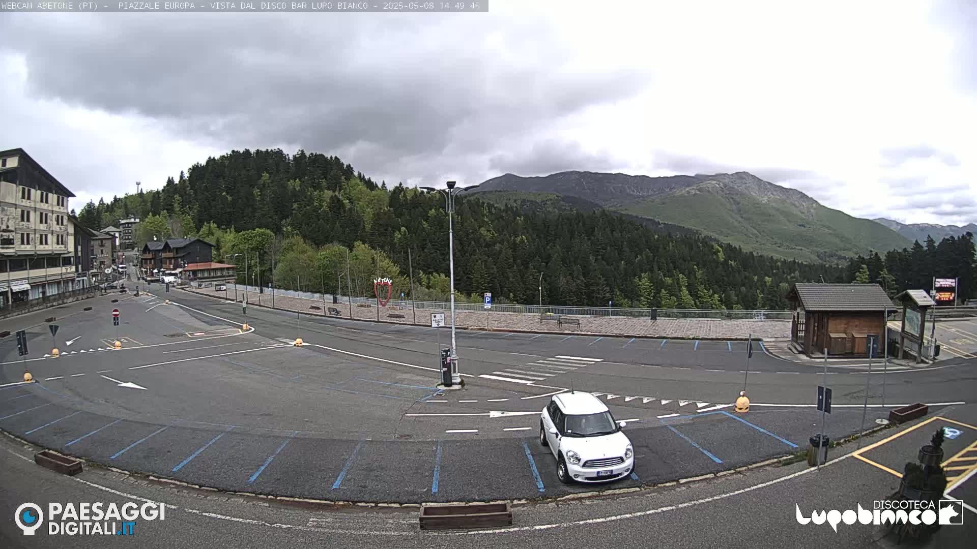 A mostly empty paved parking lot with a white SUV parked in it overlooks a town nestled in a mountain valley under a cloudy sky.