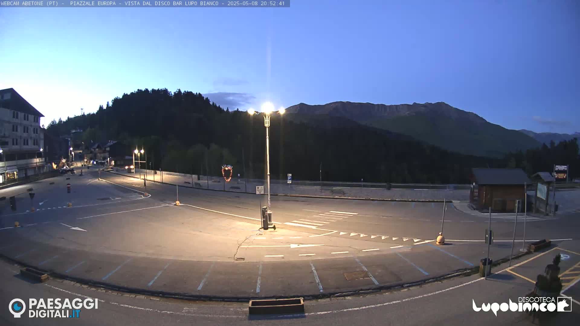 An empty parking lot and roadway are illuminated at dusk, with a mountain range in the background under a clear sky.