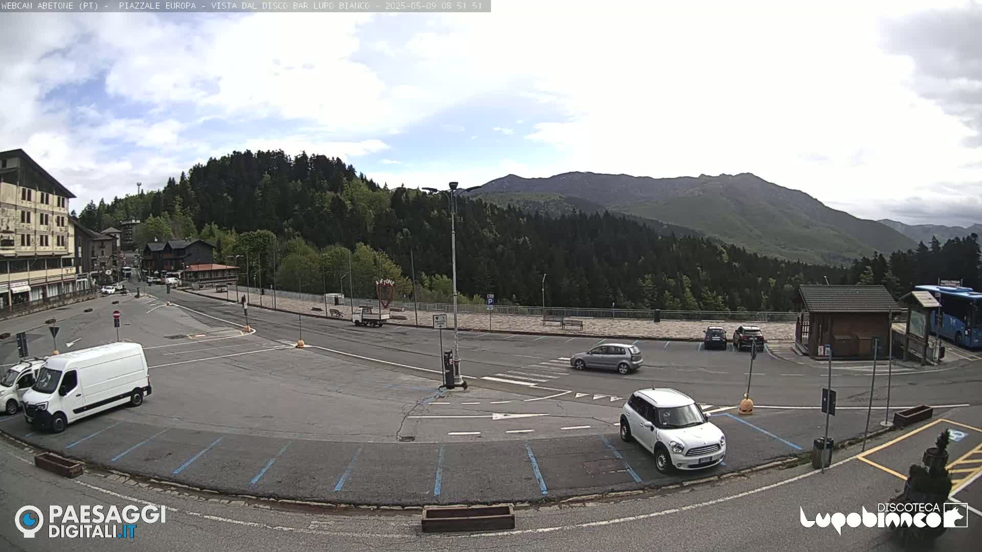 A mostly empty parking lot in a mountain town, overlooked by forested hills under a partly cloudy sky, features several cars and vans.